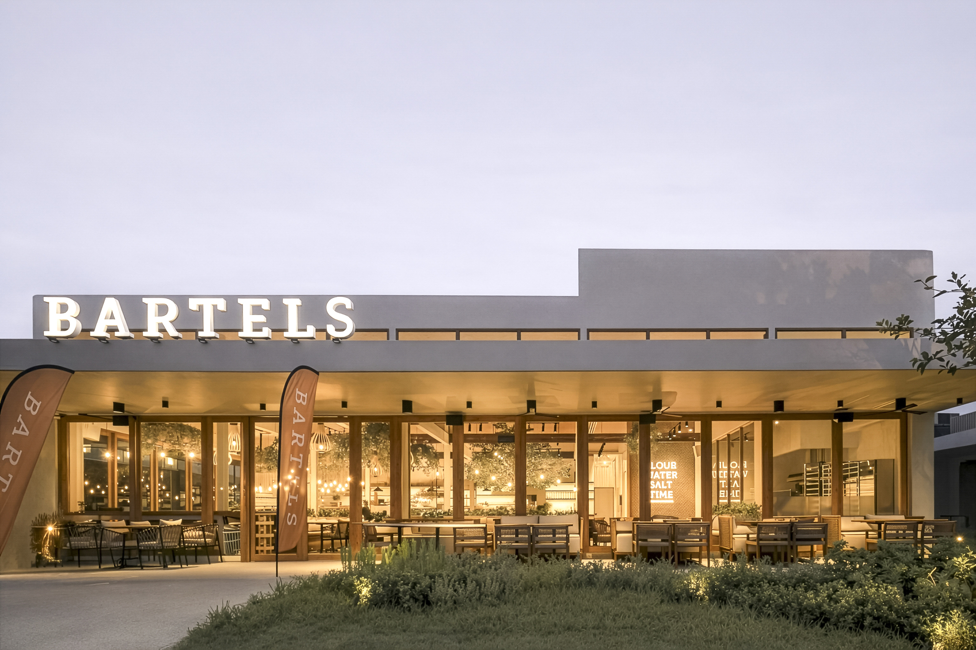Glass-walled restaurant facade with interior lighting glowing through planted forecourt at twilight