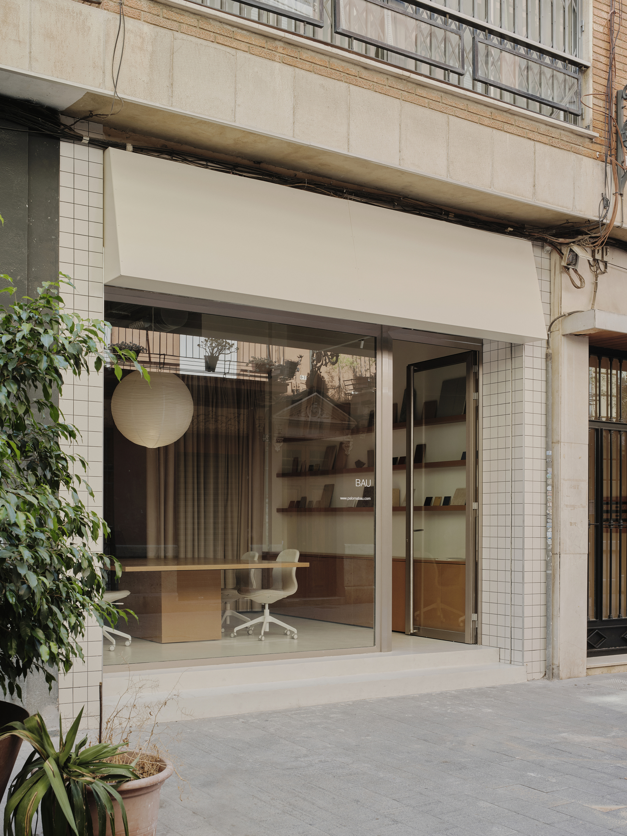 Street-facing storefront with white tile surround, retractable awning, and glazed opening revealing office interior with potted plants