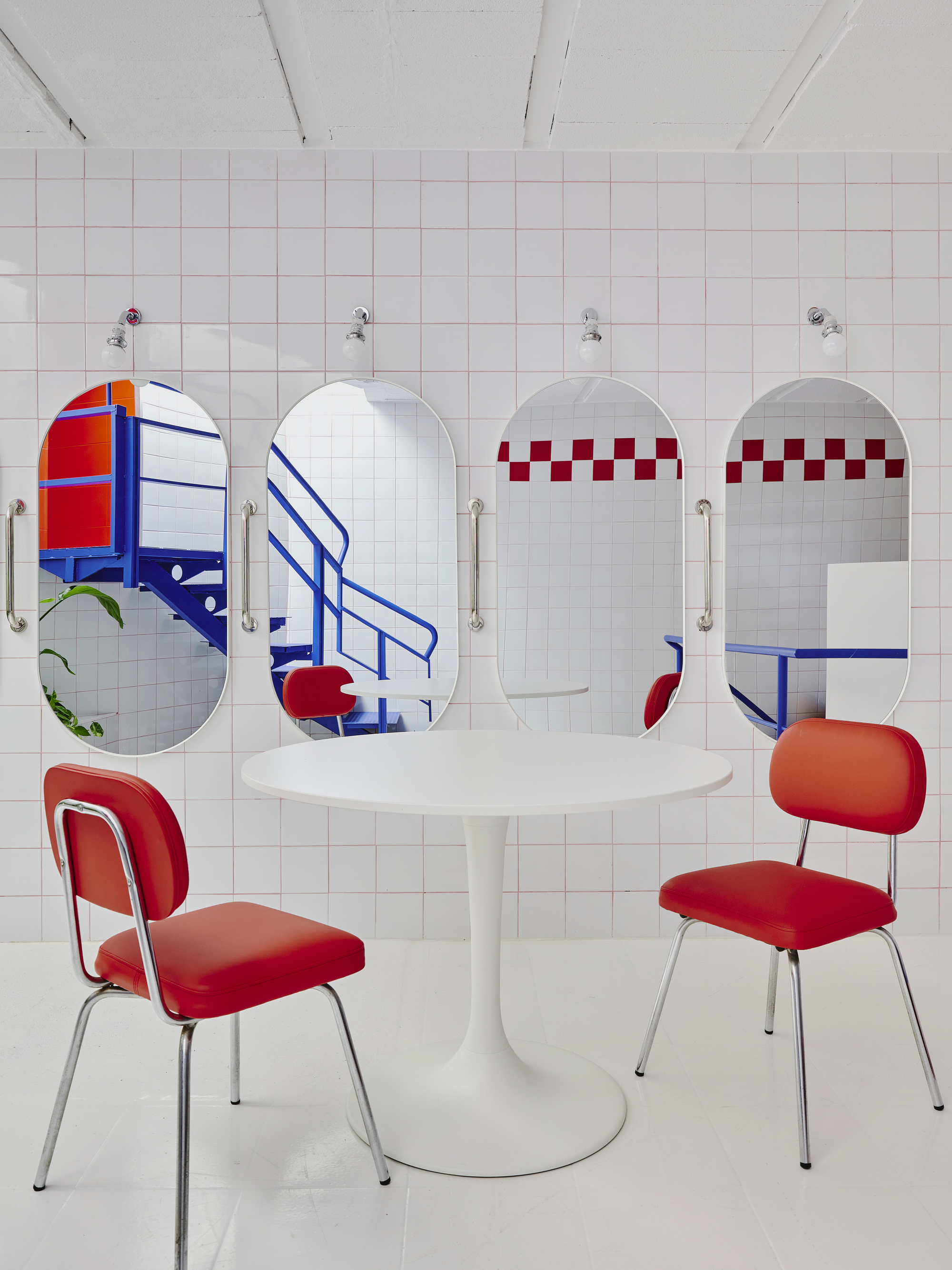 White tiled room with four oval mirrors bearing graphic patterns, white pedestal table, and red upholstered chairs