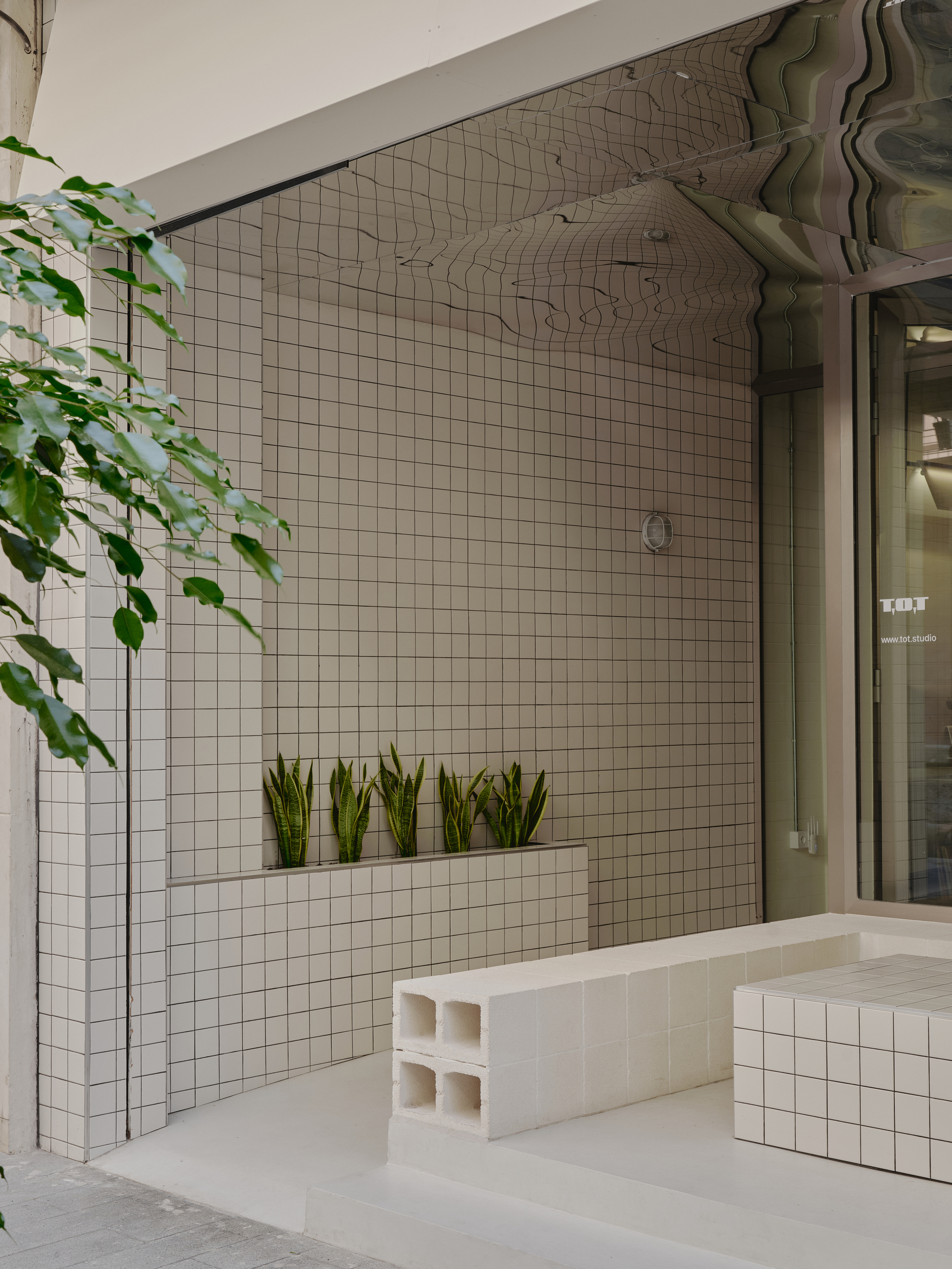 White grid-tiled courtyard wall with integrated planter shelf, trailing vines overhead, and modular tile seating blocks