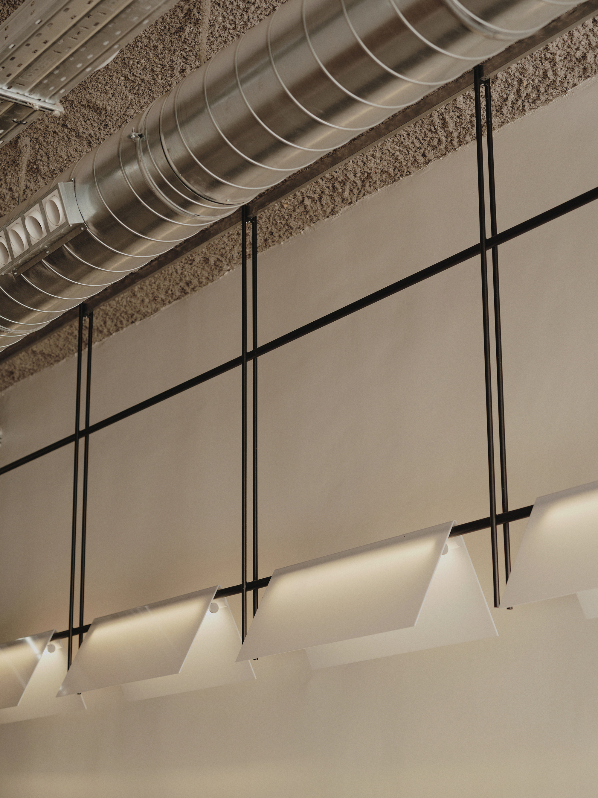 Exposed spiral ductwork running above suspended pendant lights on black cord against textured ceiling