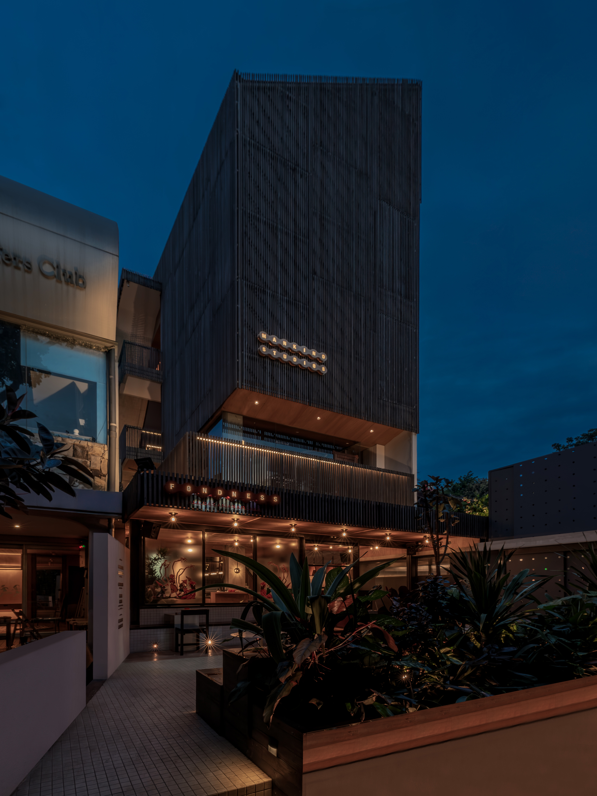 Street view of the dark timber-clad tower with illuminated signage and planted ground floor at twilight