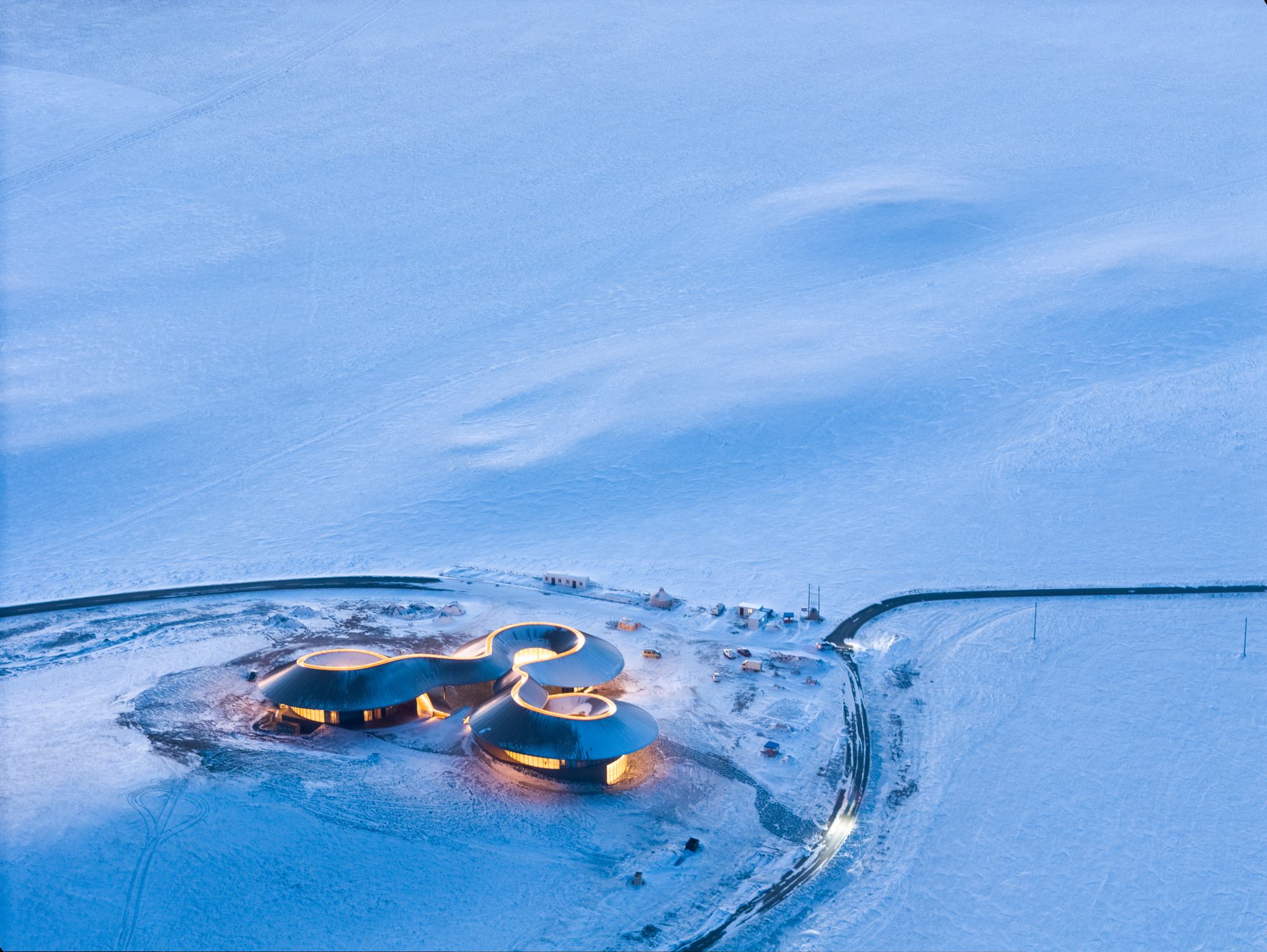 Dusk aerial with the building's interior glowing warm against the blue snow