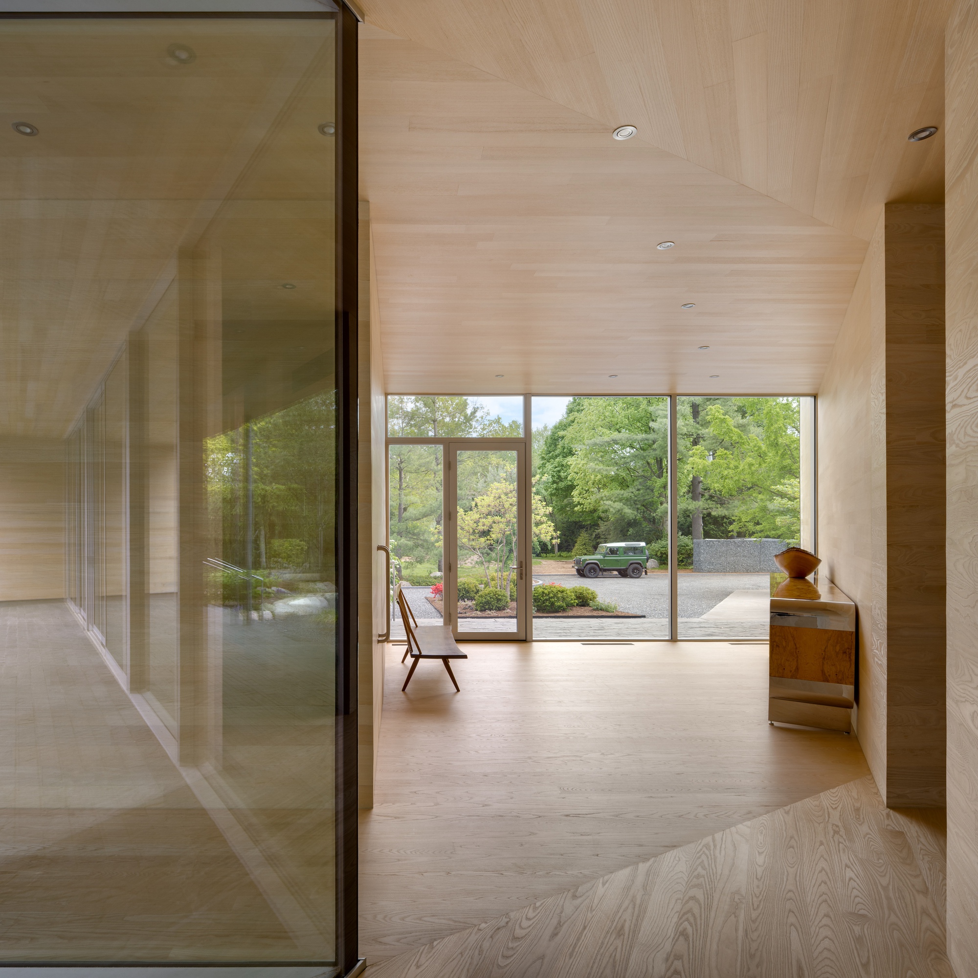 Wood-lined corridor with glass partition reflecting view through glazed doors to landscaped driveway