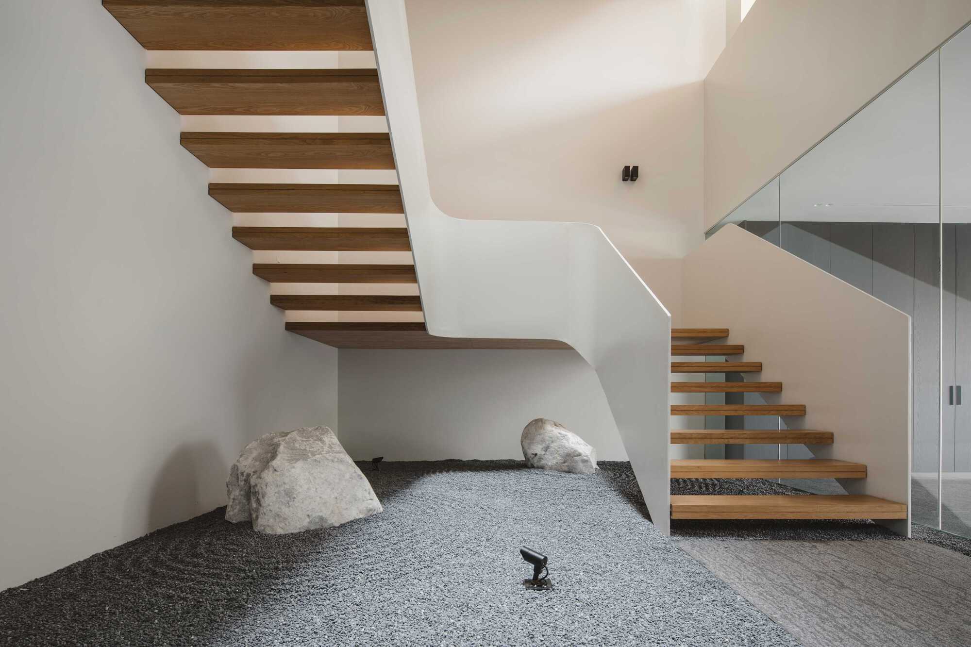 Floating timber staircase wrapping around a pebbled interior courtyard with white sculptural walls and skylight above