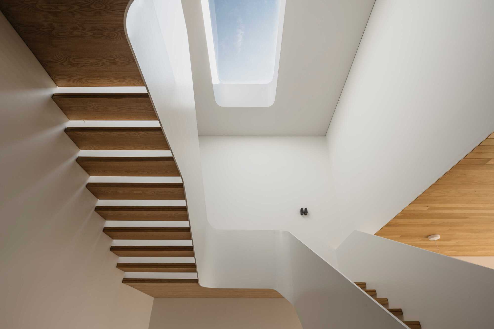 View up the cantilevered timber stairwell toward a rectangular skylight framed by white walls