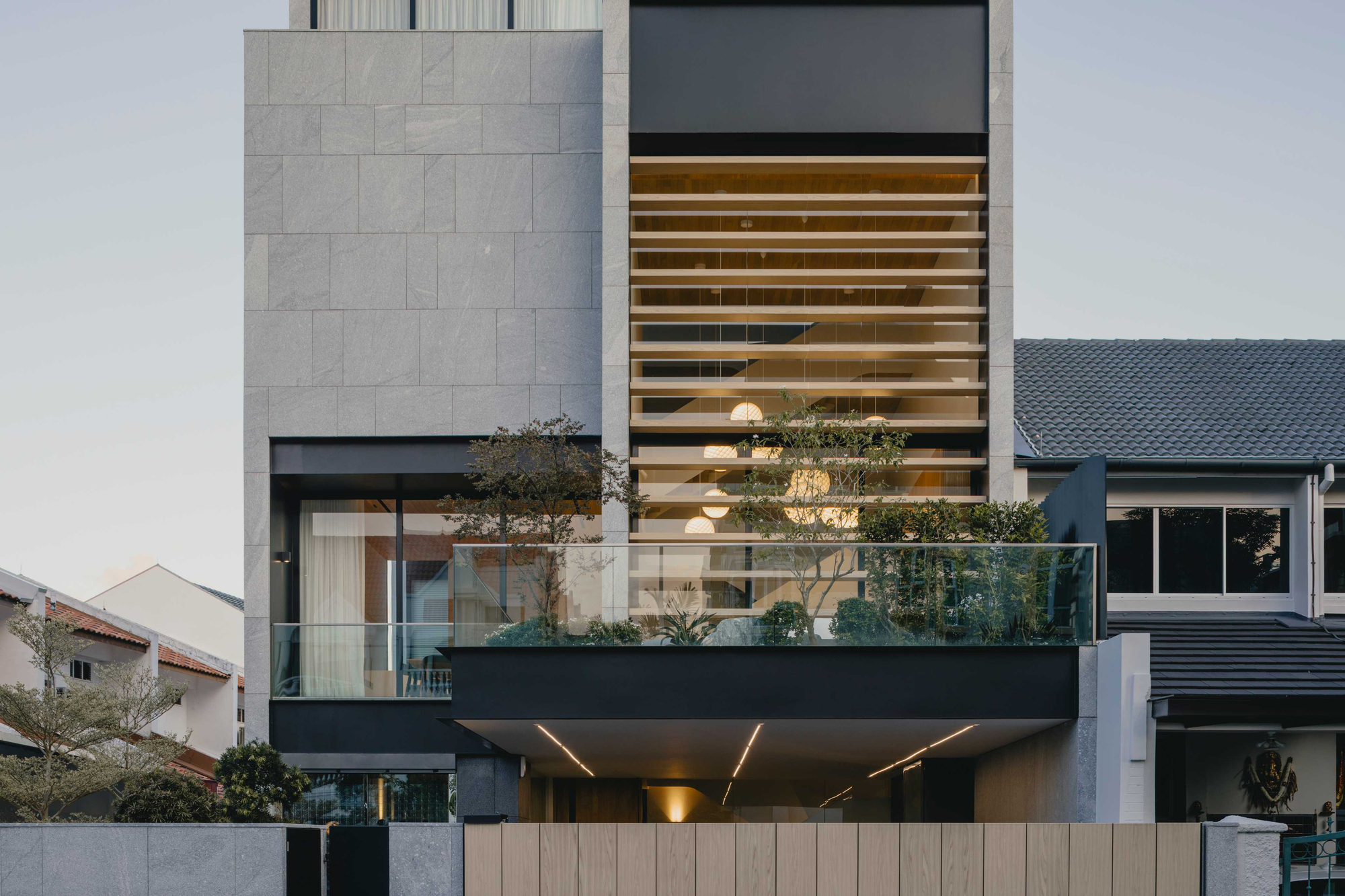 Street view of the grey stone facade with horizontal timber screening and a planted balcony at dusk