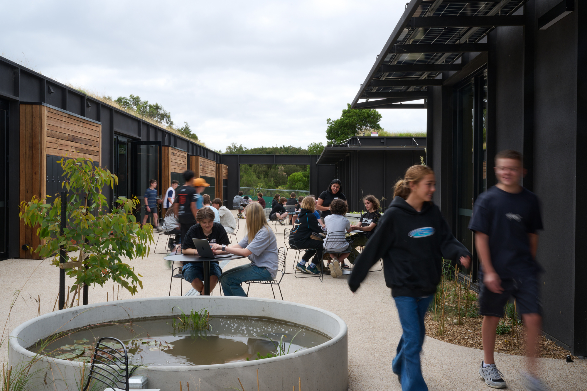 Central courtyard at lunch with students at outdoor tables, a round concrete water feature, and photovoltaic canopies above