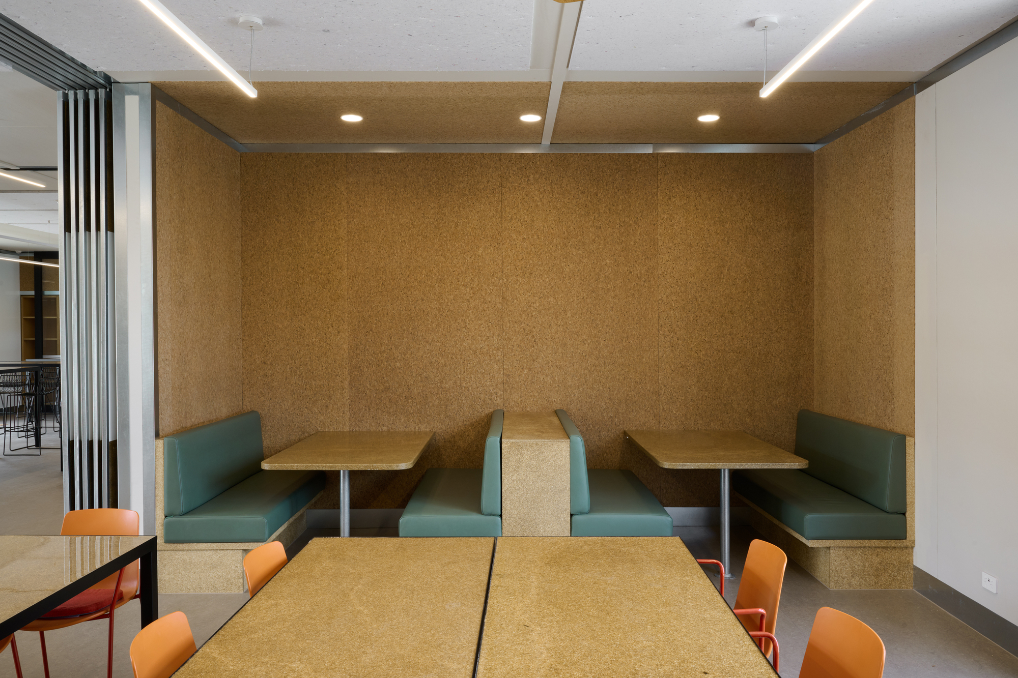 Interior booth seating nook with cork walls, teal banquettes and cork-topped tables under linear lights