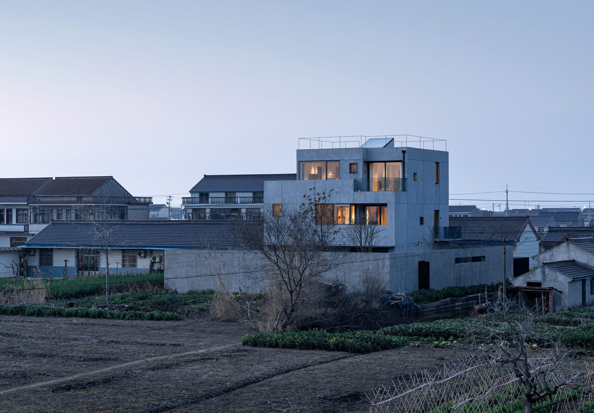 White concrete house with rooftop terrace rising above neighboring buildings and cultivated fields at dusk