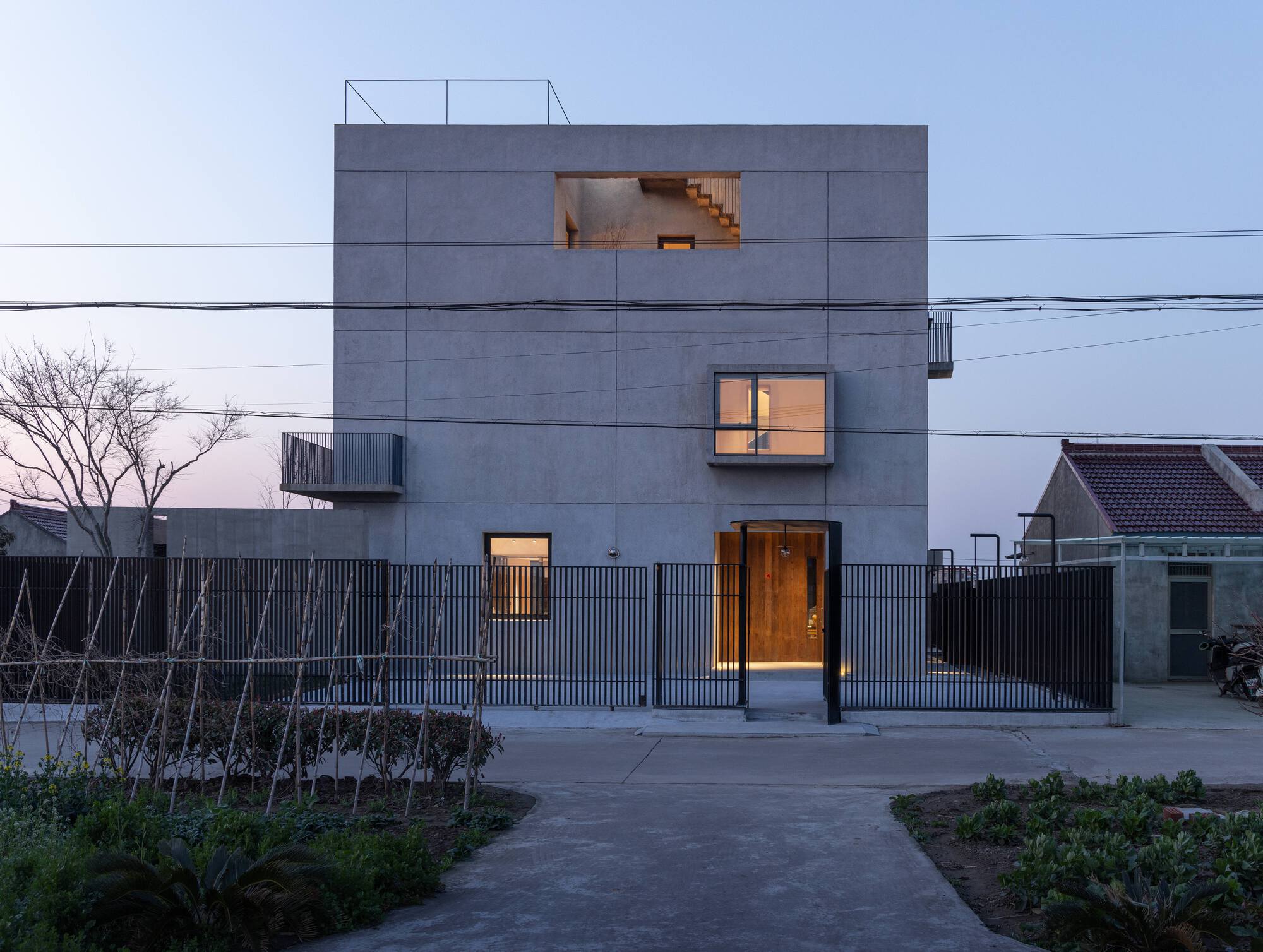 Cubic concrete volume with illuminated openings behind a slatted metal fence and bare tree at dusk