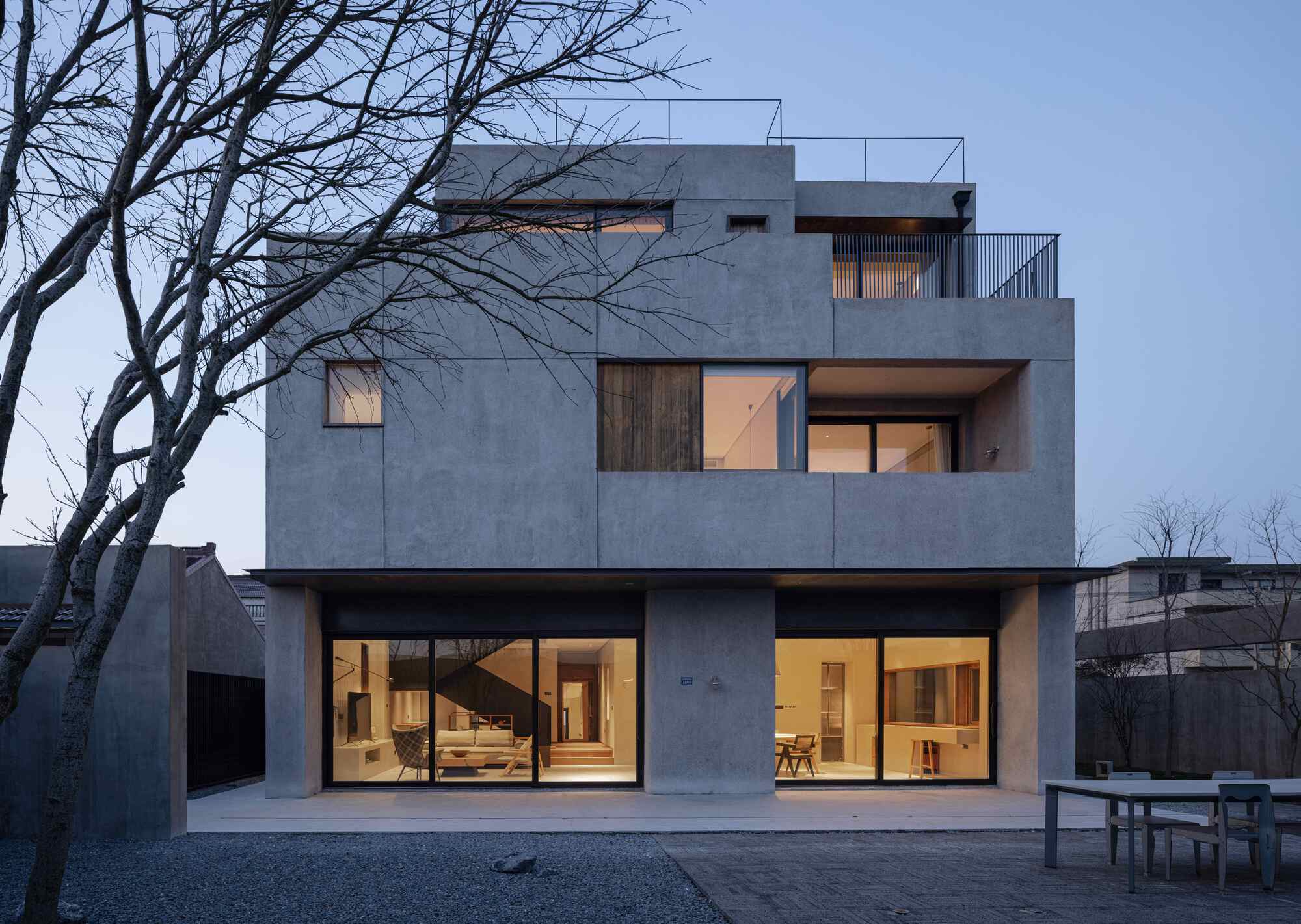 Rear elevation with large glazed openings and cantilevered terraces against a gravel courtyard at twilight