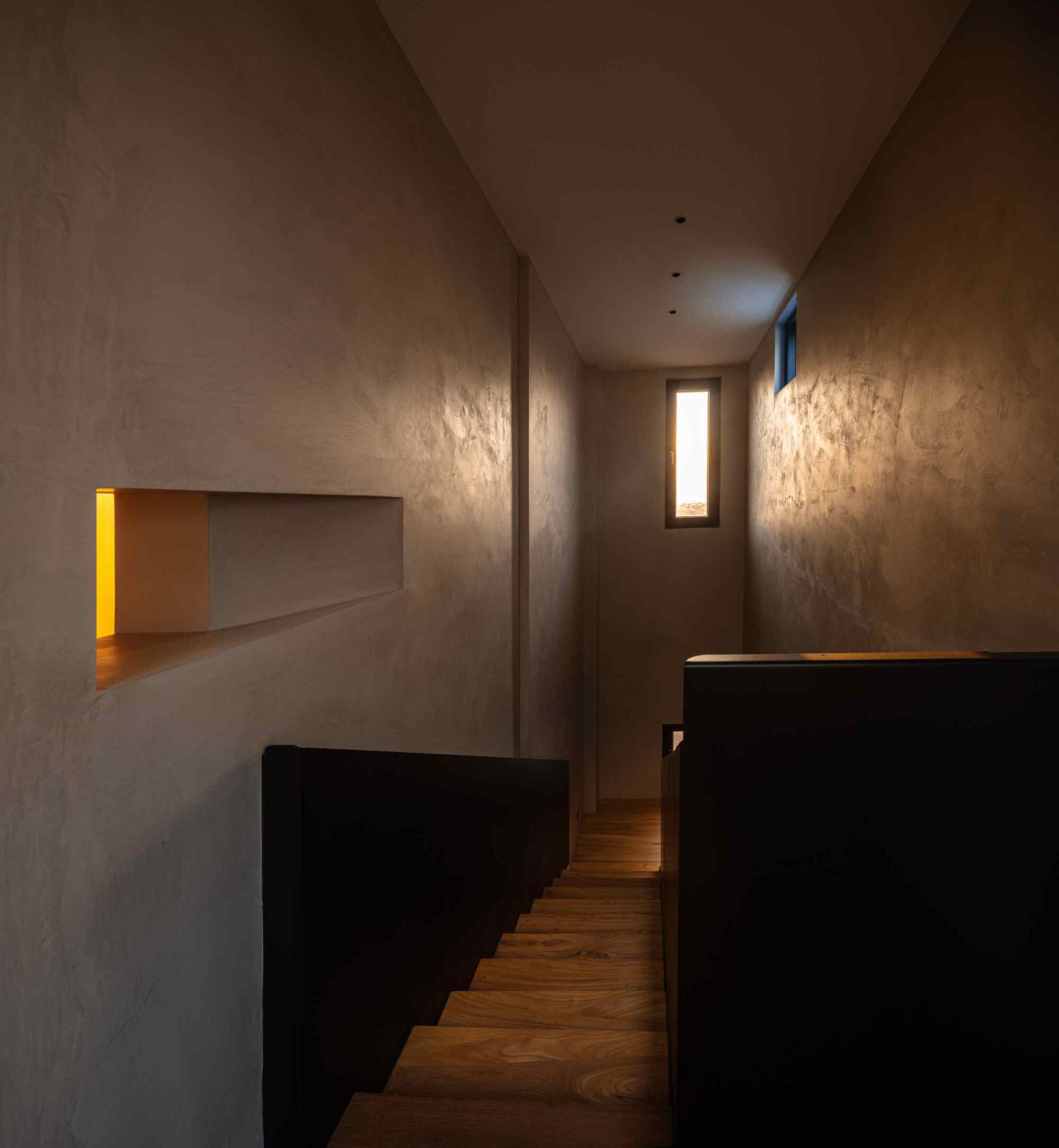 Narrow stairwell with black metal sides, timber treads and narrow window slits creating dramatic light patterns