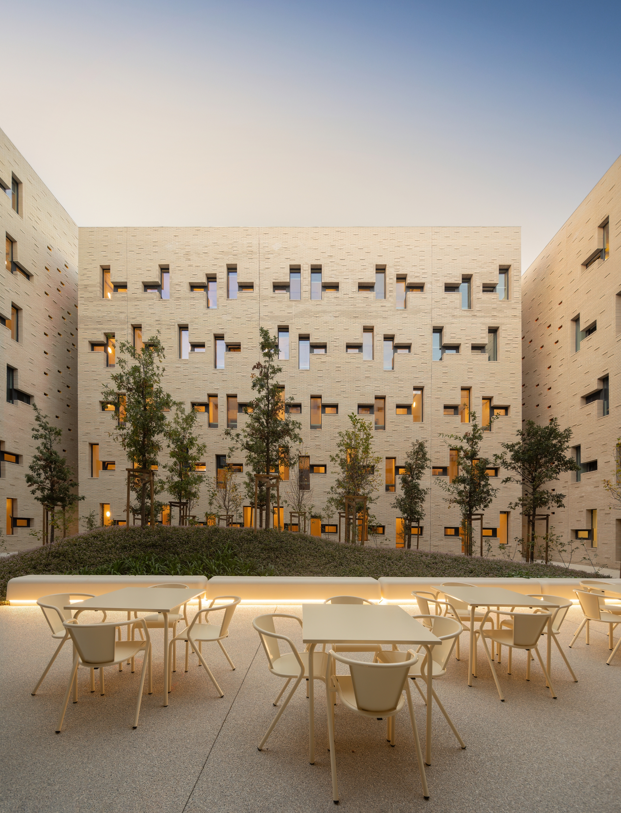 Outdoor seating area with tables and chairs facing planted trees at dusk