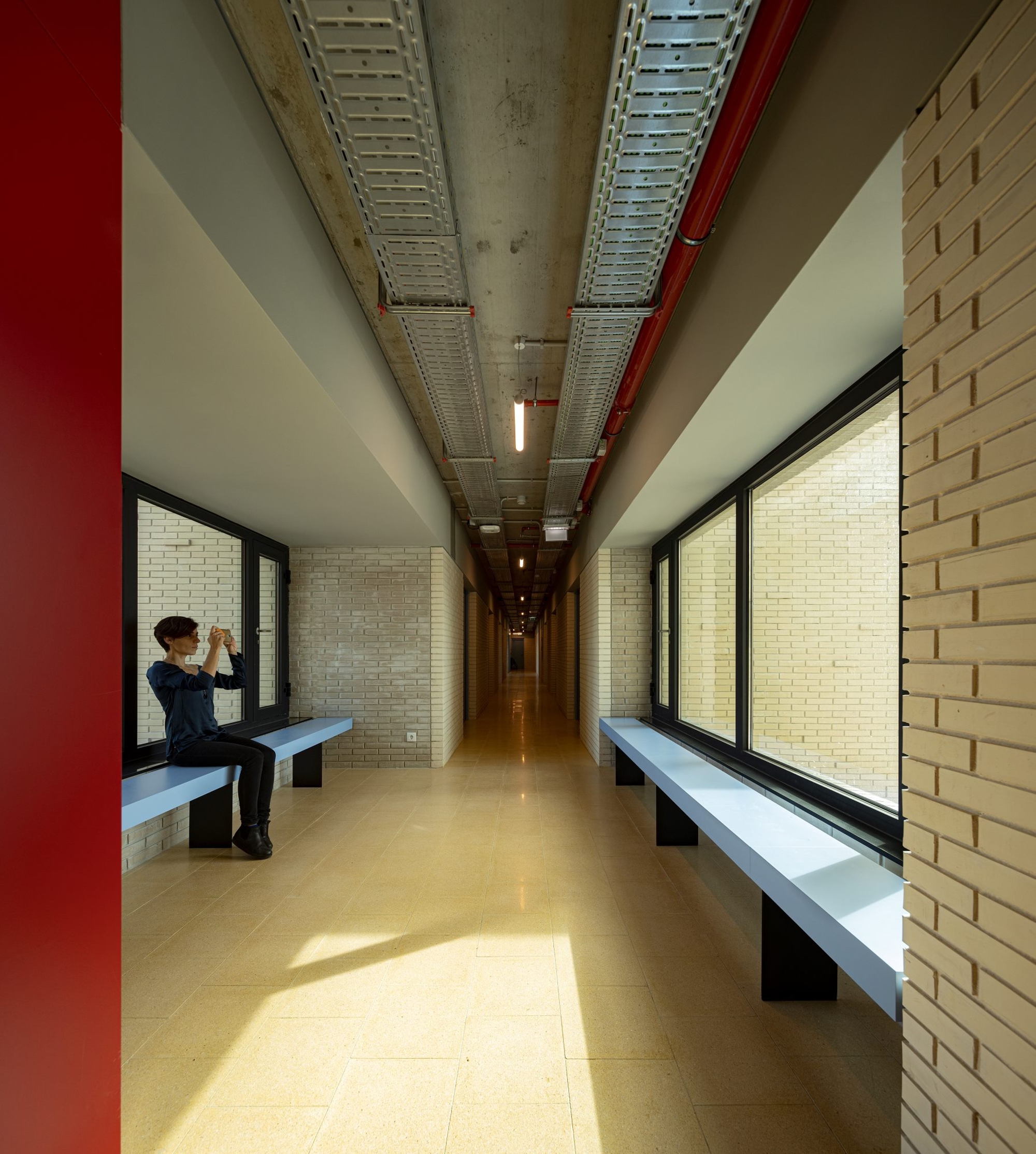 Long corridor with brick walls, bench seating, and exposed ceiling ducts with a seated figure