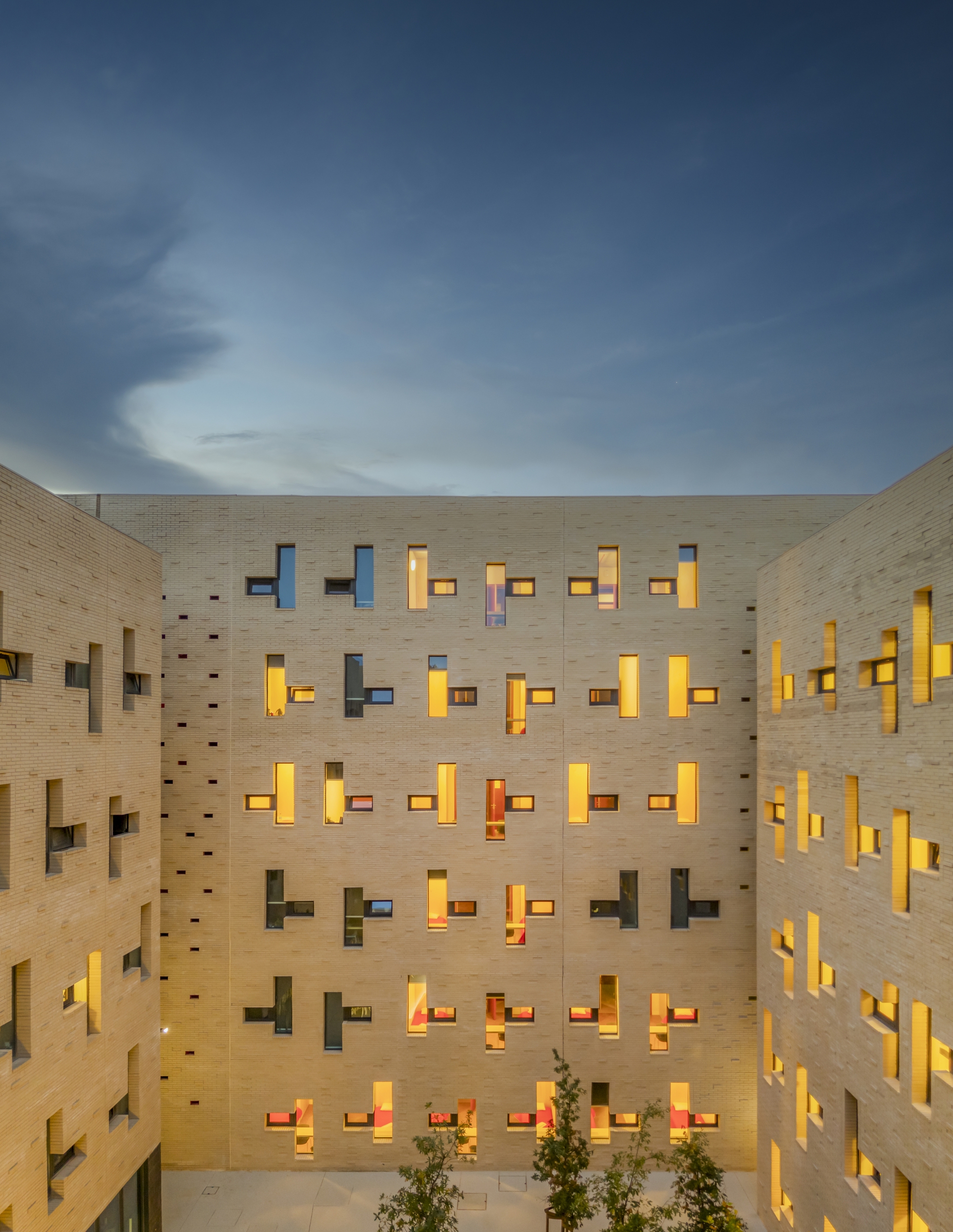 Interior courtyard surrounded by brick walls with illuminated staggered windows and small trees at twilight