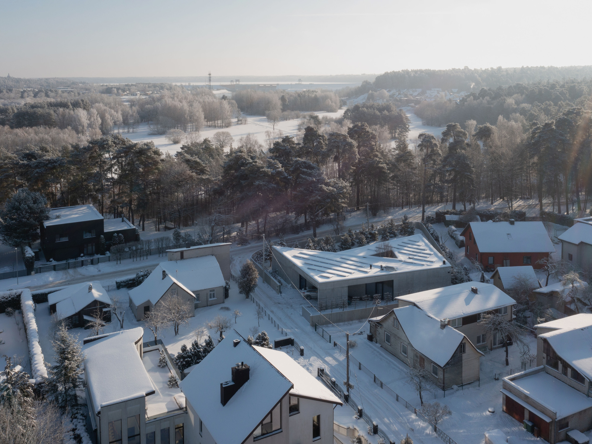 Aerial perspective of the angular roof with parallel skylights surrounded by residential rooftops and a forest in winter