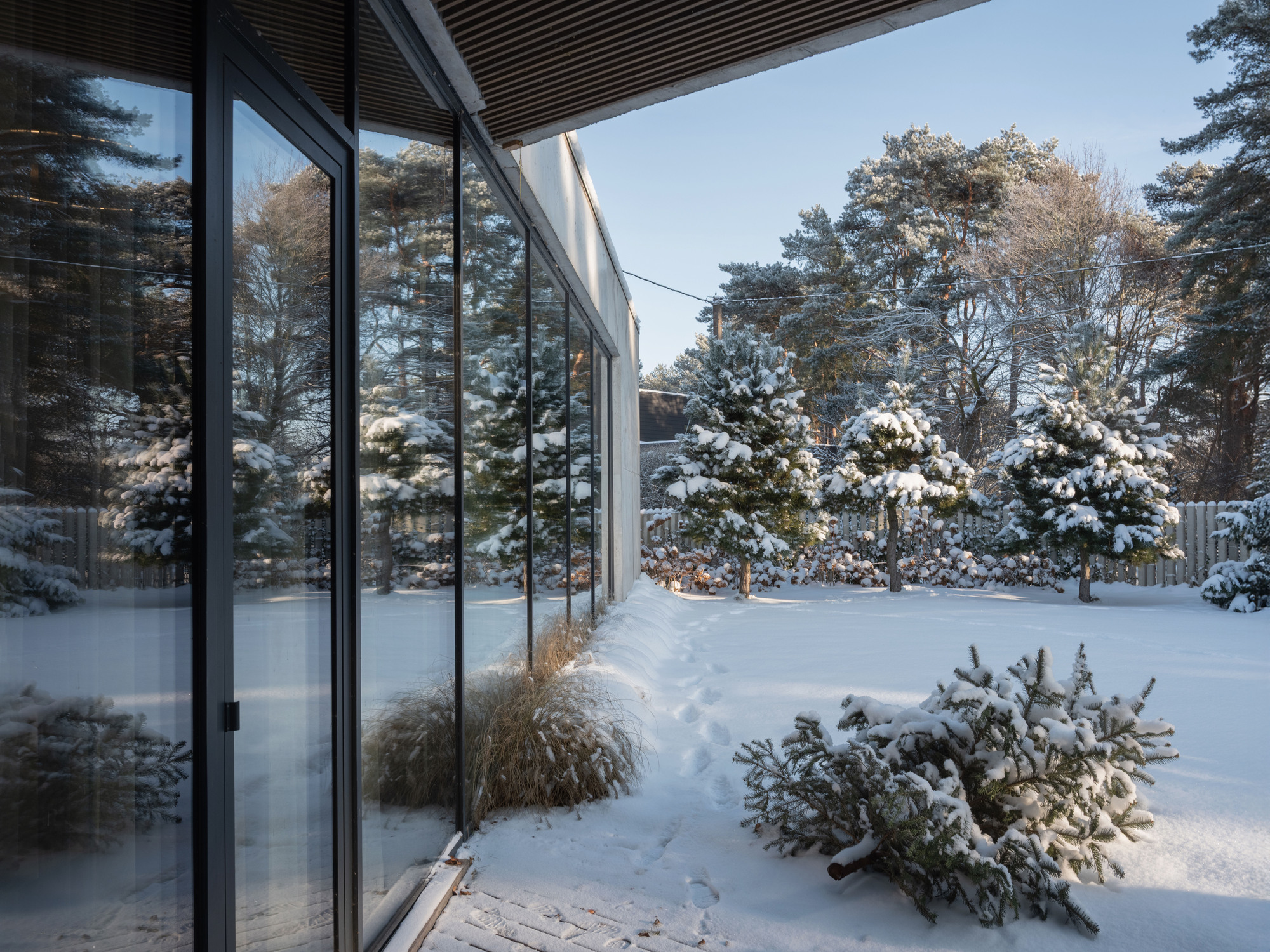 Glass facade with dark frames overlooking a snow-covered garden with planted beds and evergreen shrubs