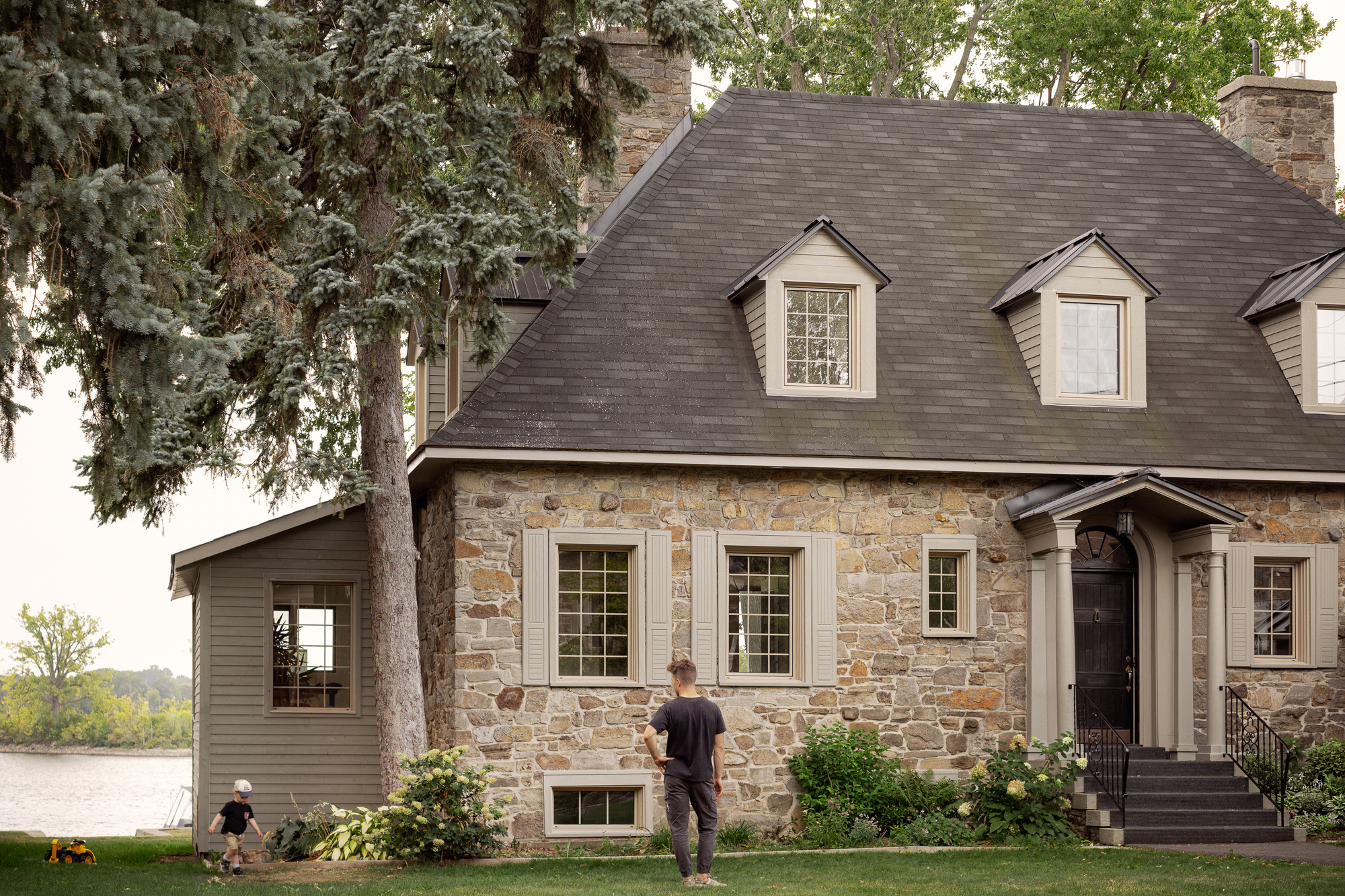 Stone facade with dormer windows and steeply pitched slate roof beside a lawn with figures and waterside trees