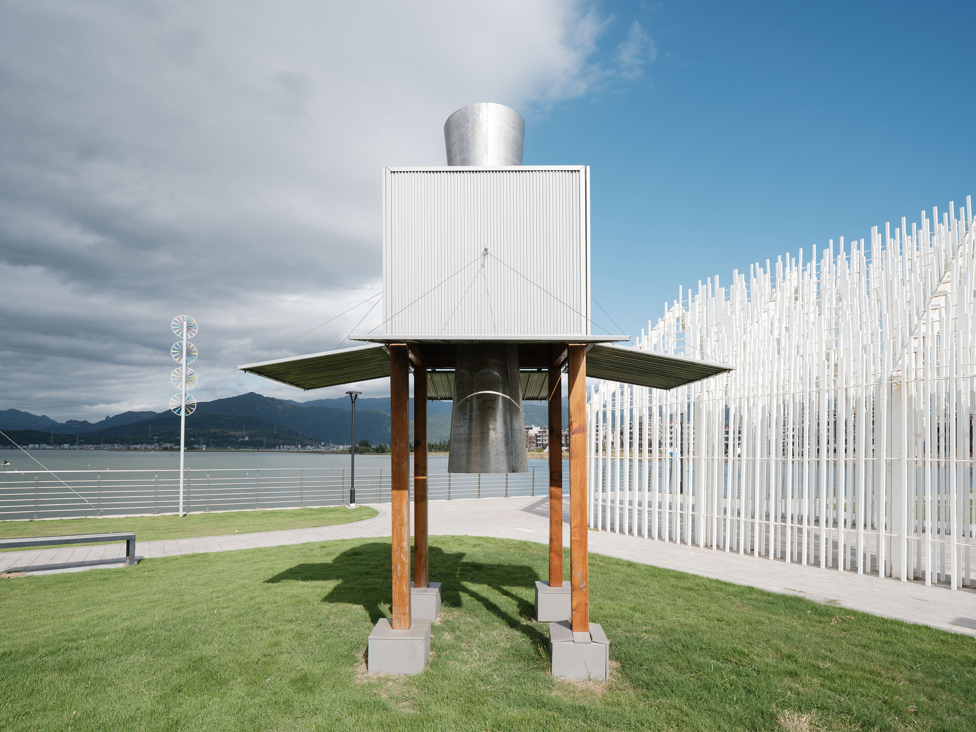 White cubic volume elevated on timber columns with a flat roof beside a vertical rod sculpture
