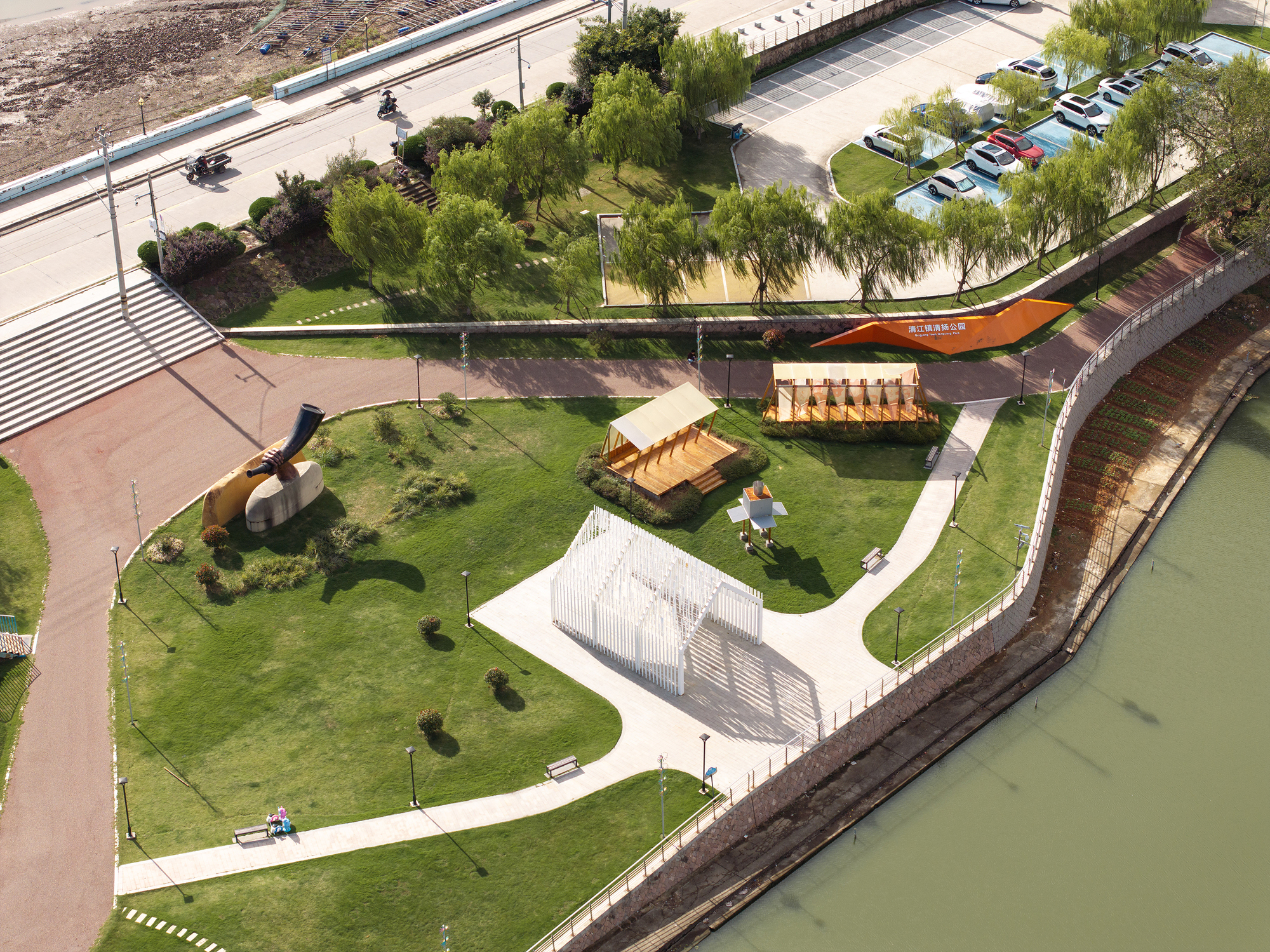 Aerial perspective of waterside installations including timber-clad structures and white vertical-screen pavilion beside the paved path