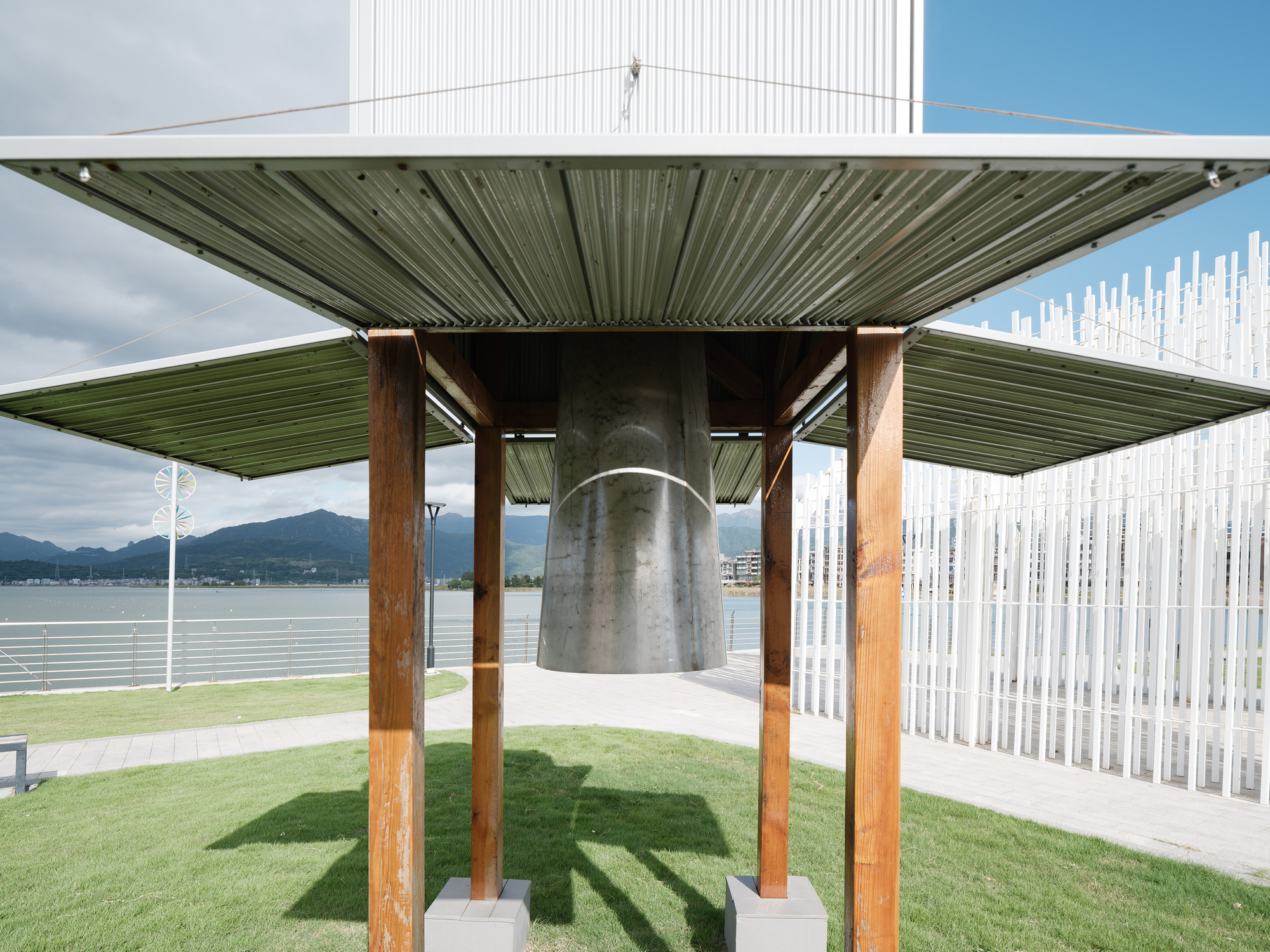 View beneath the metal canopy supported by timber columns, with suspended bell and white vertical screen beyond