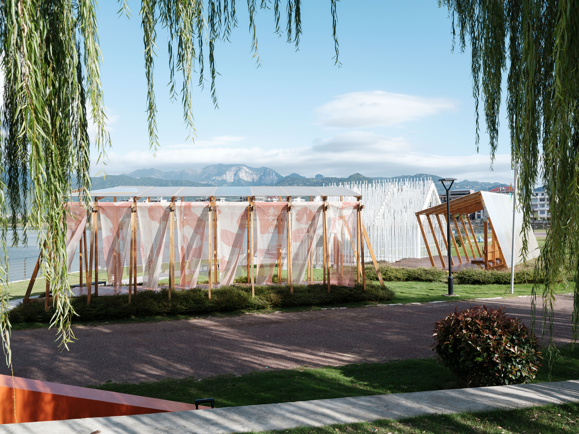 Timber frame pavilion with pink translucent fabric panels framed by willow branches along a lakeside path