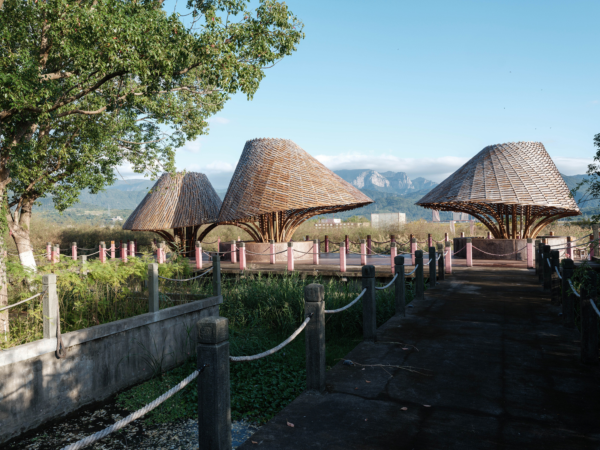 Three thatched bamboo canopies with branching timber columns along a boardwalk with distant mountains visible