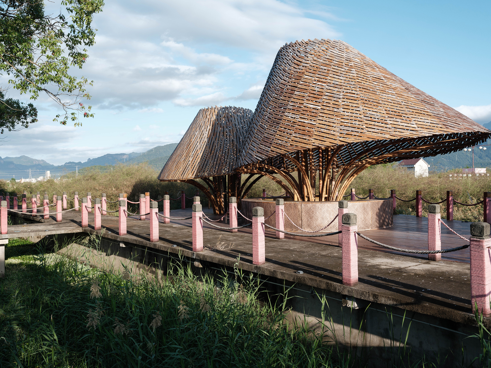 Two layered bamboo roofs supported by splayed timber columns beside a boardwalk lined with pink posts