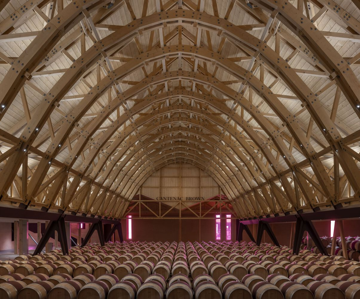 Interior view of arched timber trusses spanning above rows of wine barrels with pink accent lighting