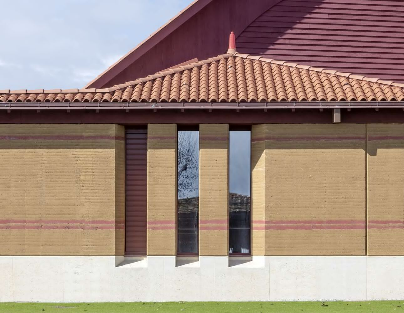 Exterior wall with rammed earth banding, vertical window slits and clay tile roof under blue sky