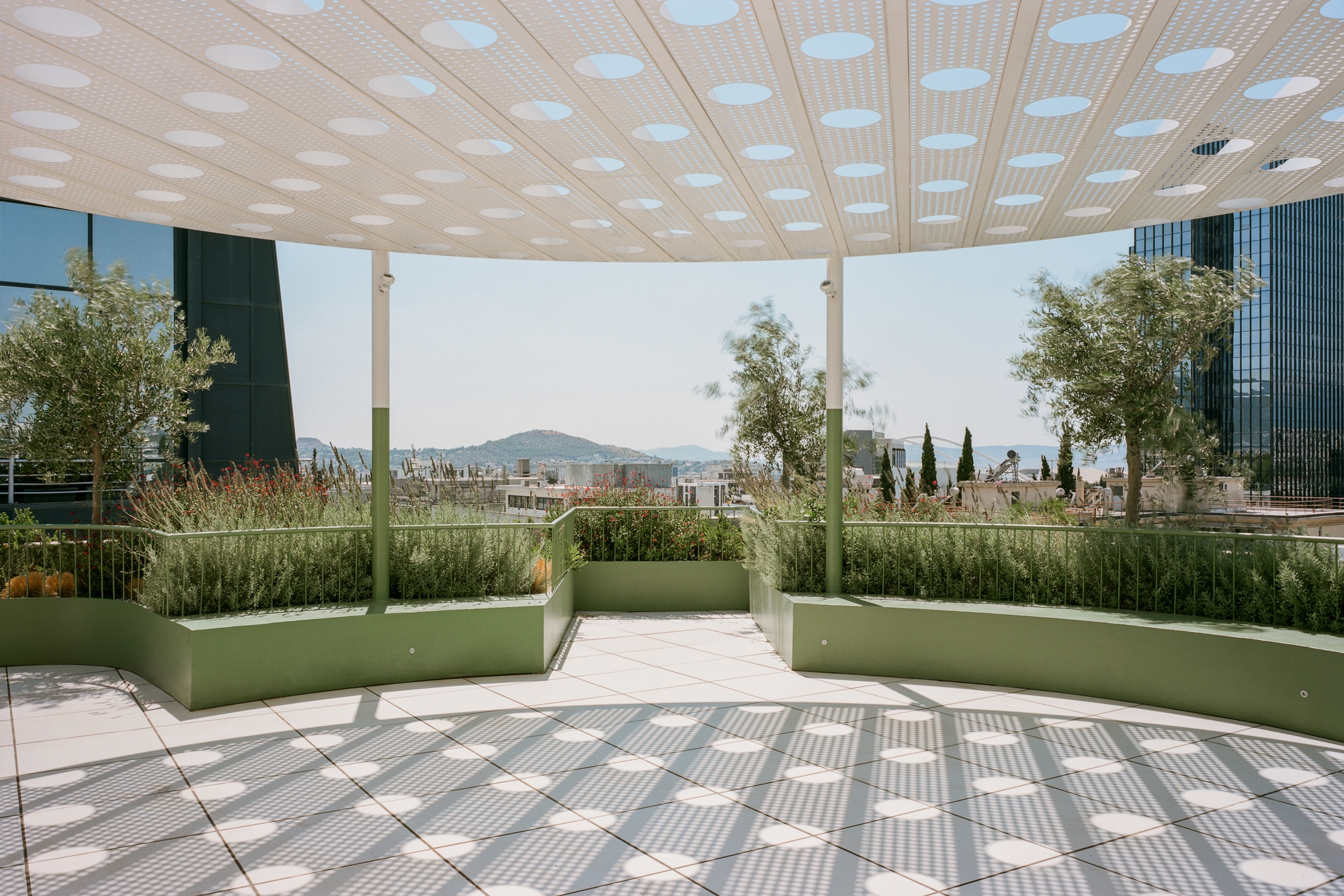 Circular canopy with perforated ceiling above green planted beds and tiled terrace overlooking distant hills