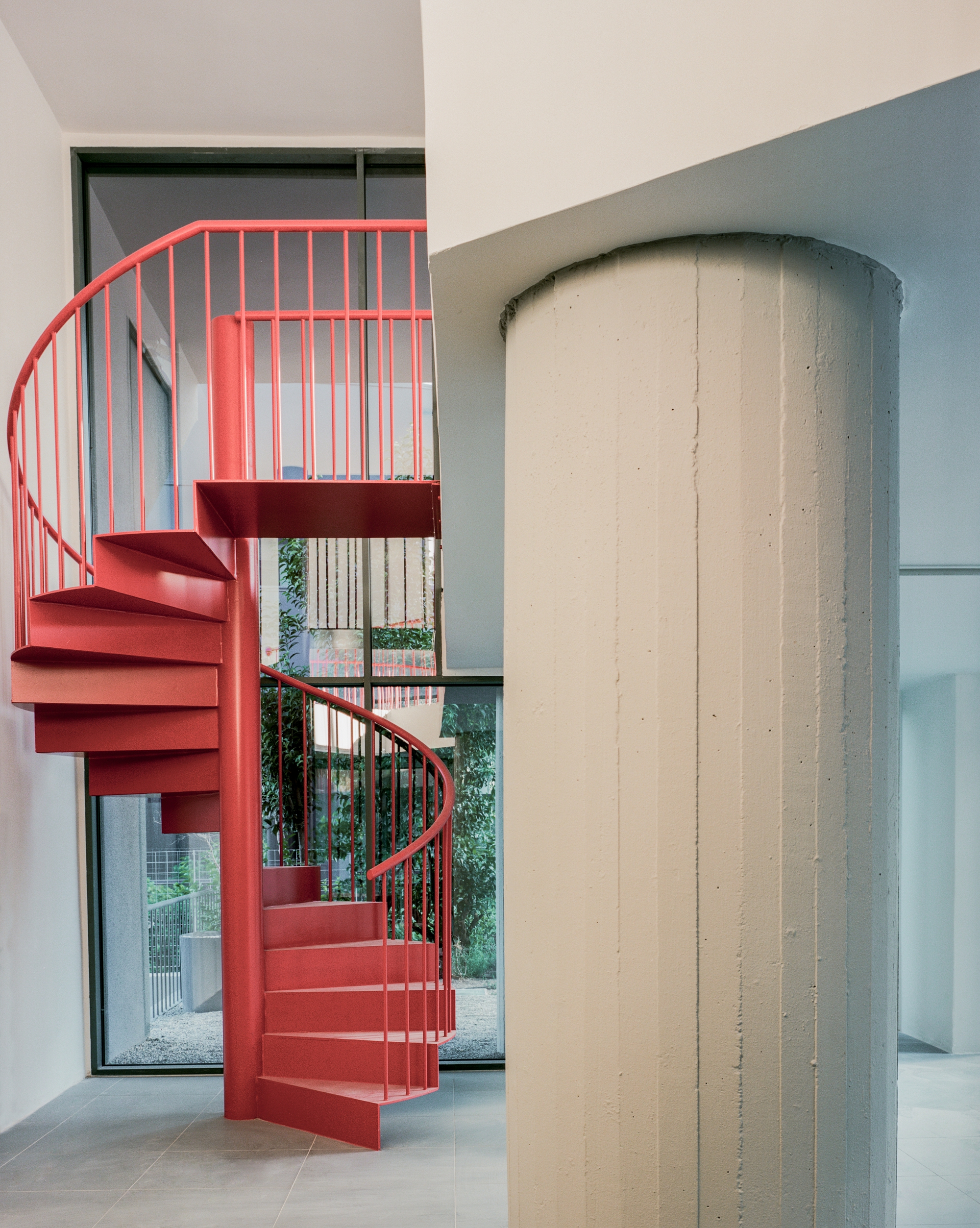 Red spiral staircase beside a cylindrical concrete column with garden views through glazed opening