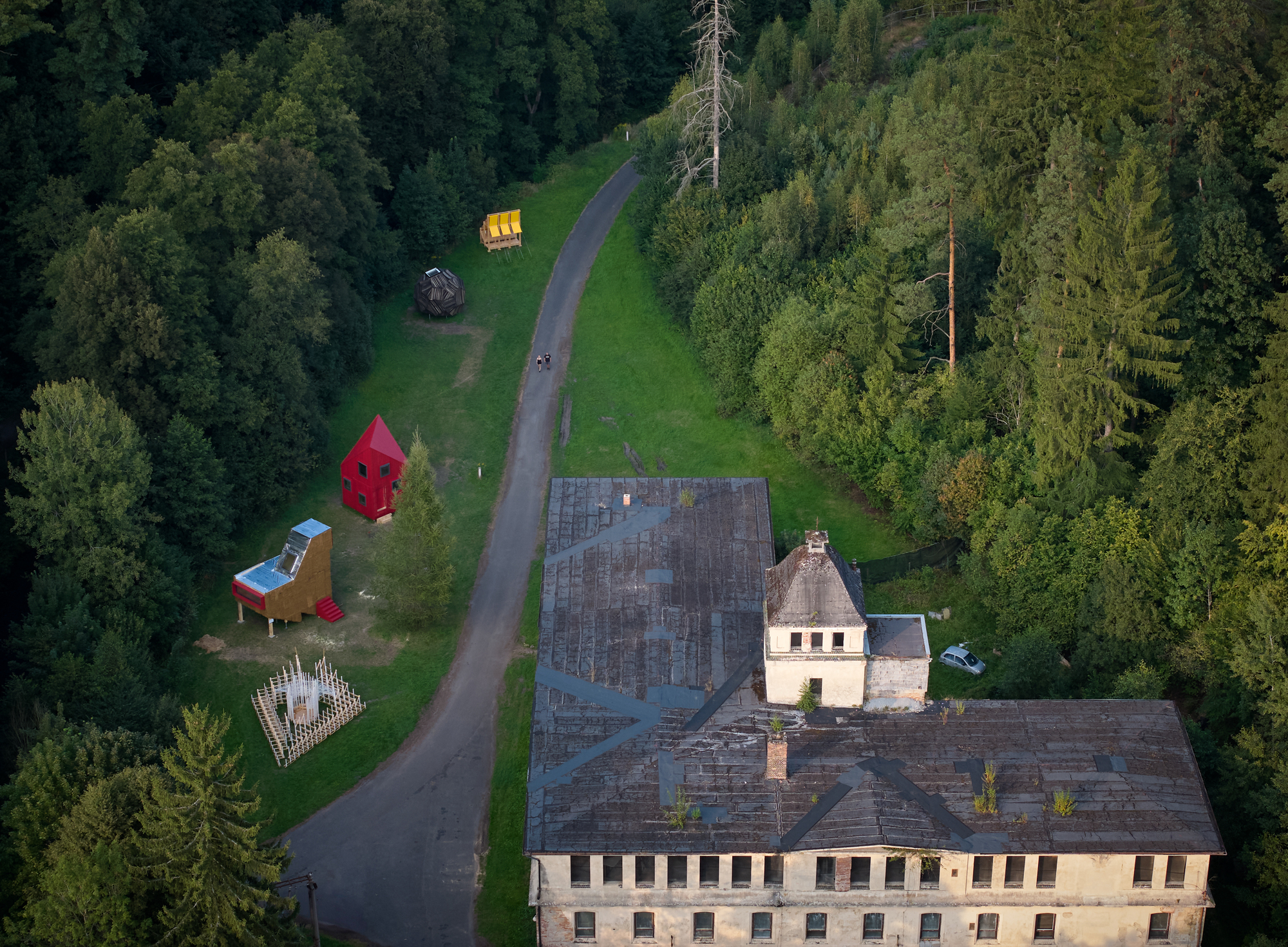 Aerial view of sculptural pavilions scattered across a parkland beside a weathered institutional building