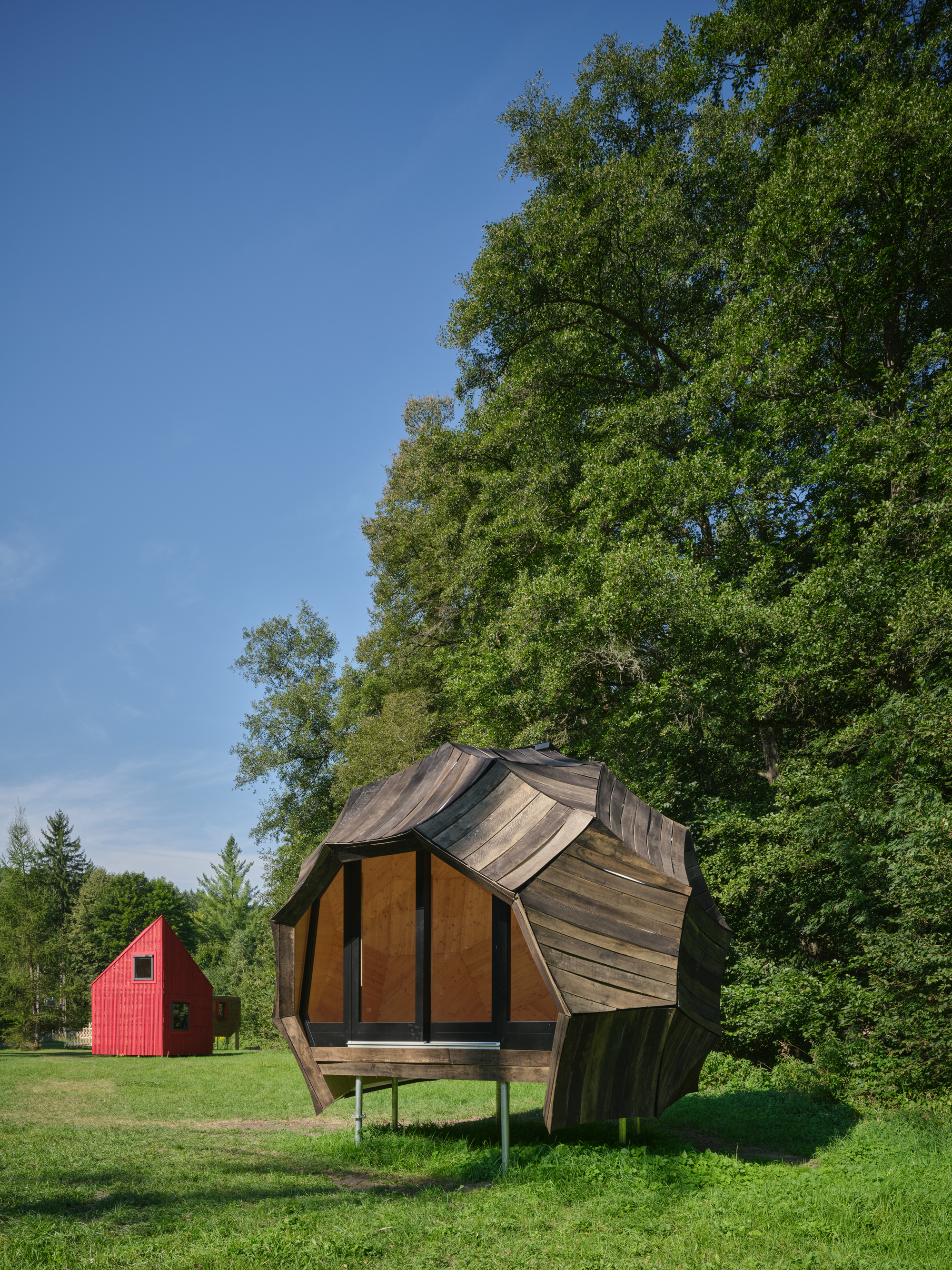 Faceted timber pavilion on stilts with angular roof planes beneath tall deciduous trees