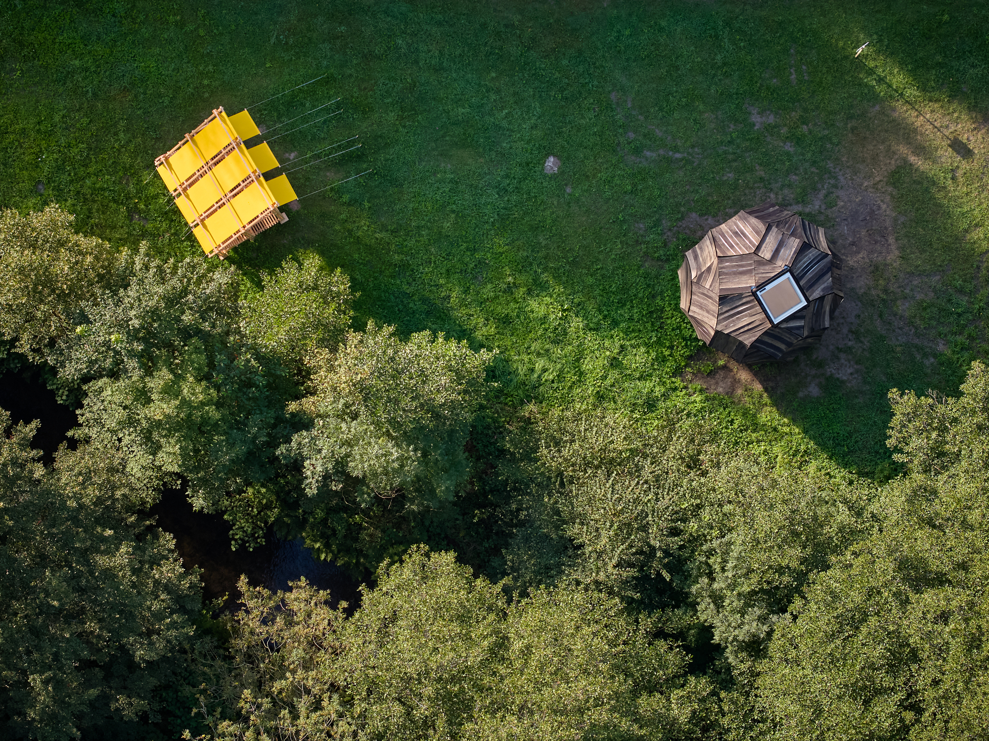 Drone view of yellow pavilion and faceted timber structure on a clearing edged by forest