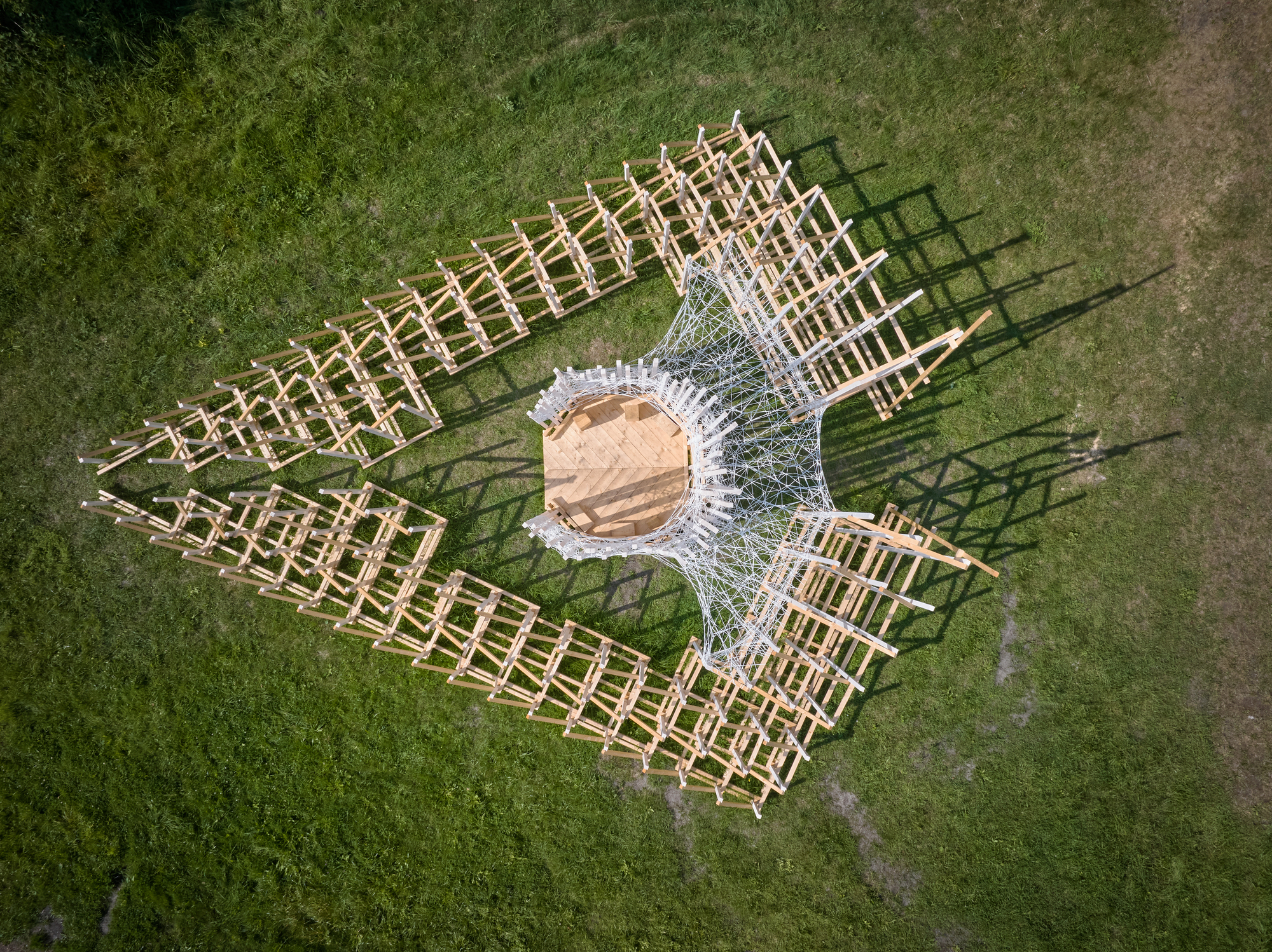 Aerial view of a timber lattice structure with central rope tensioning on a grassy field