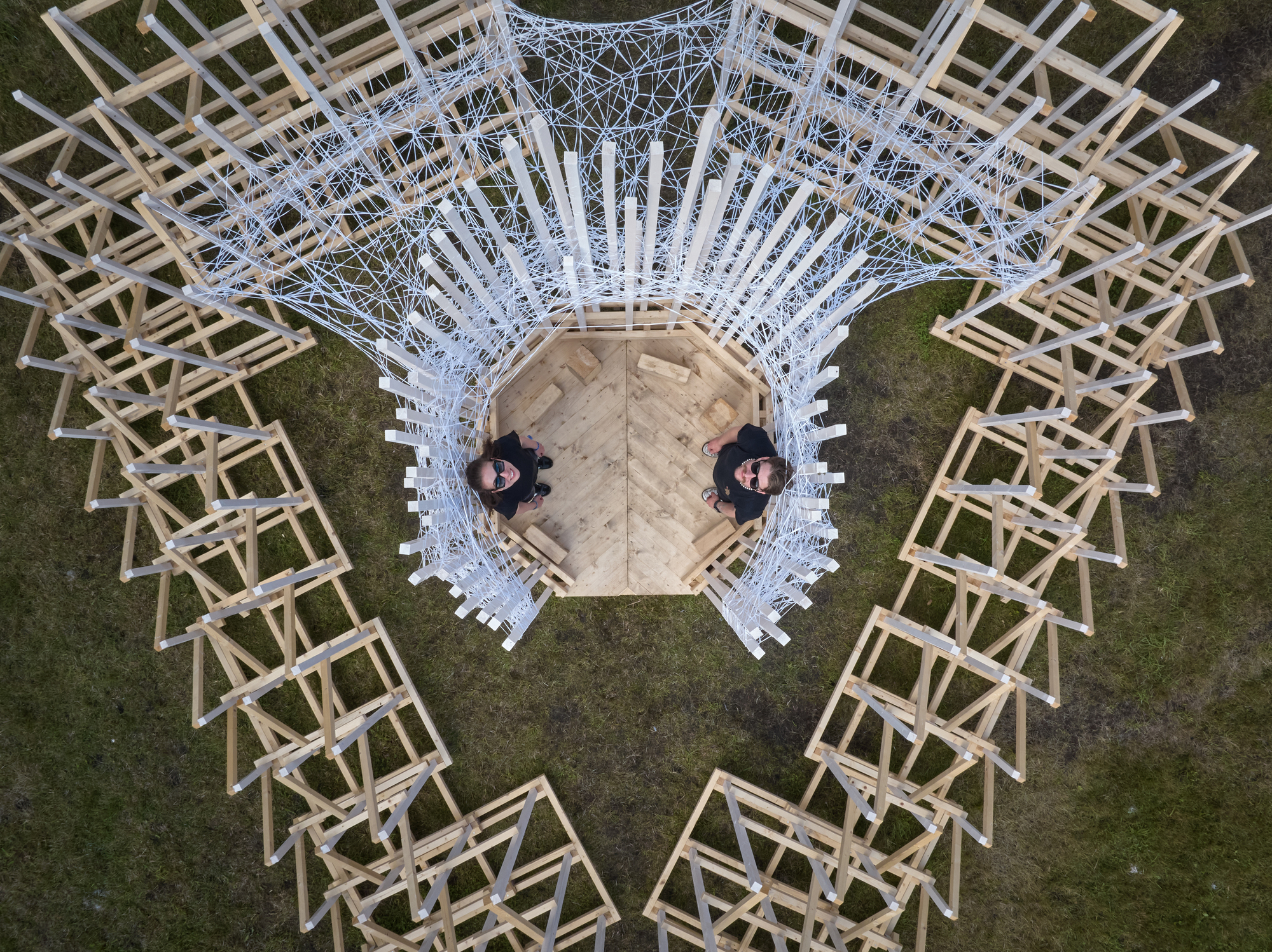 Aerial view of a radial timber pavilion with white lattice canopy and two people standing on the central platform