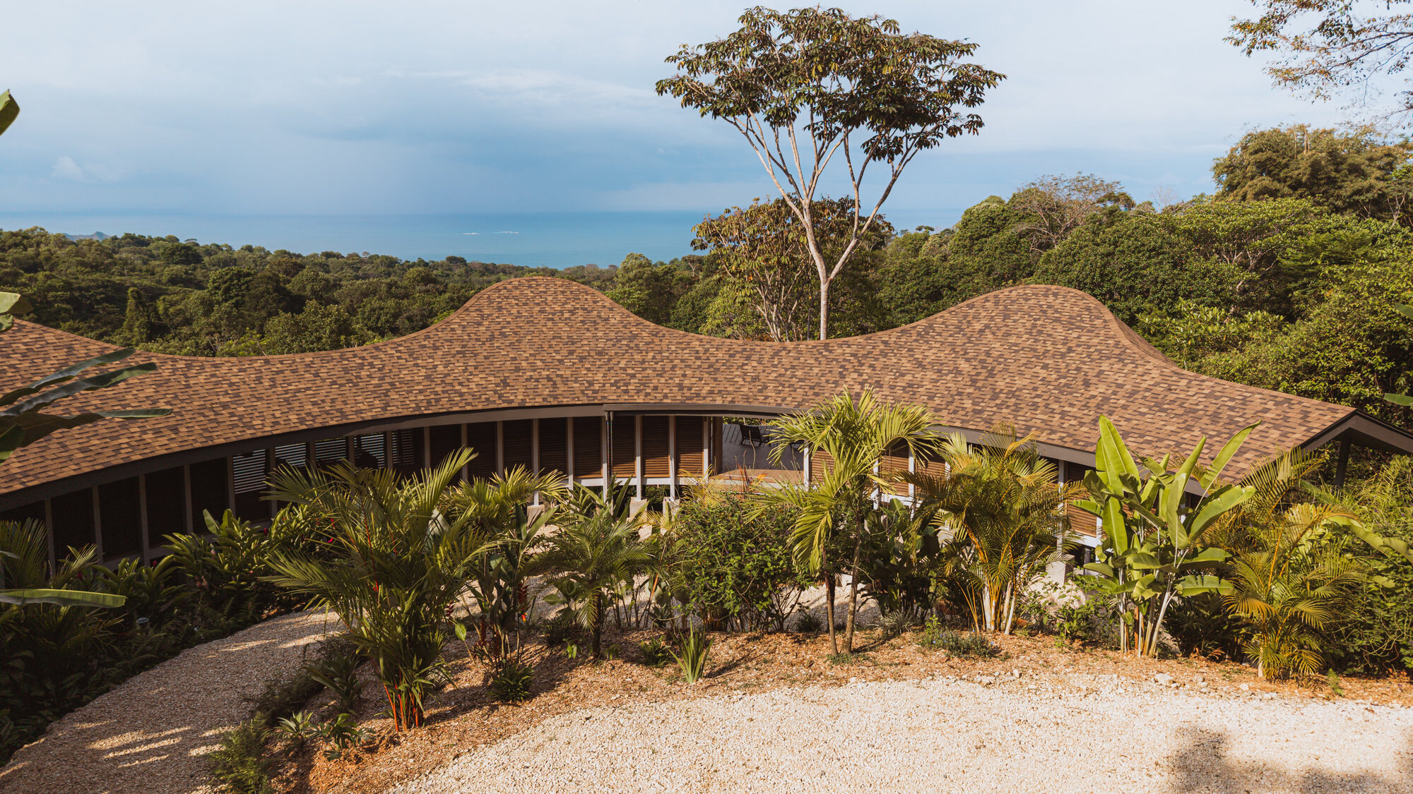 Front view of the undulating shingle roof stretching horizontally above the tropical garden with the Pacific Ocean in the distance