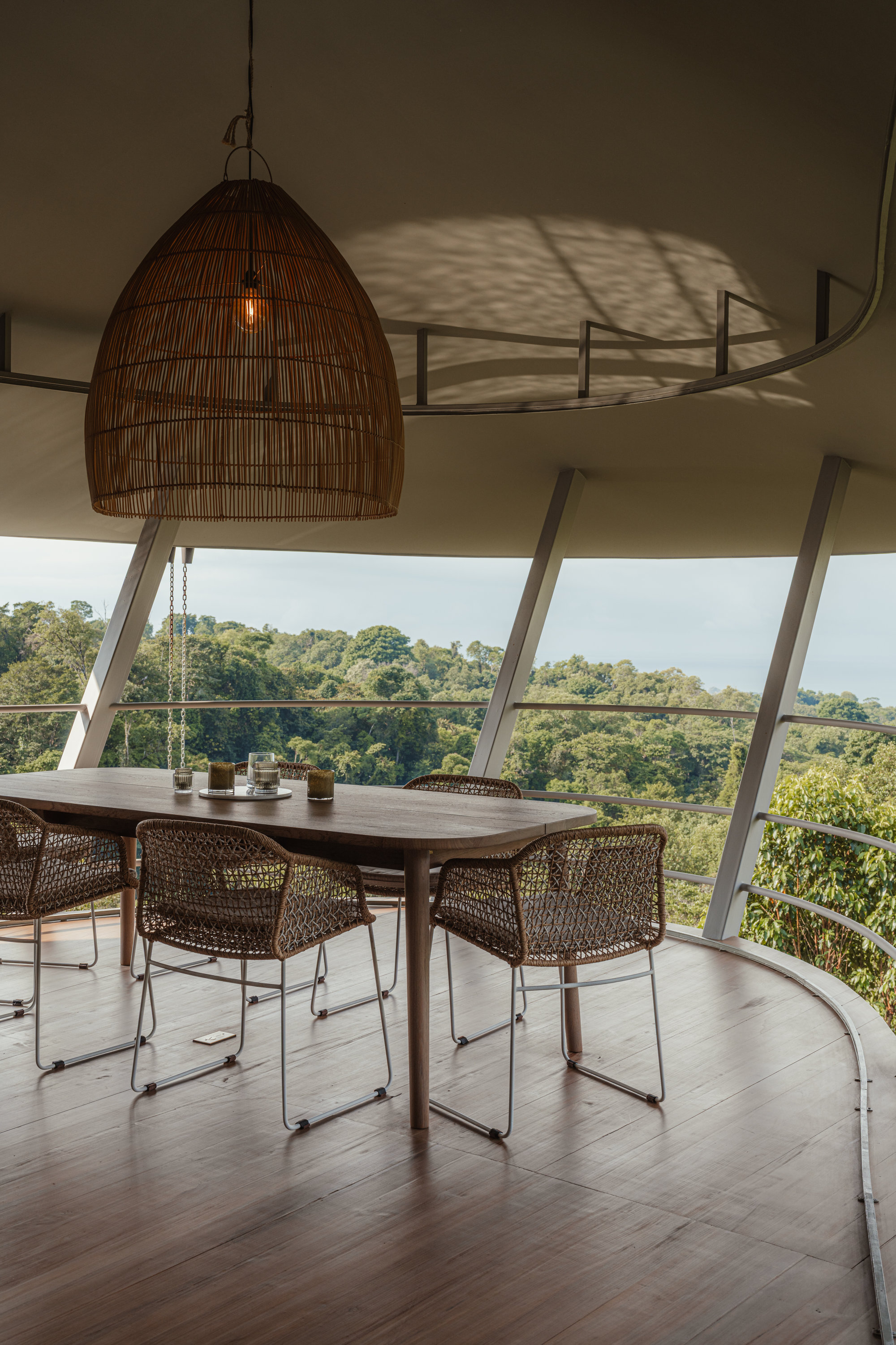 Dining area inside the curved volume with a large woven pendant light and views out to the forest through the glass perimeter