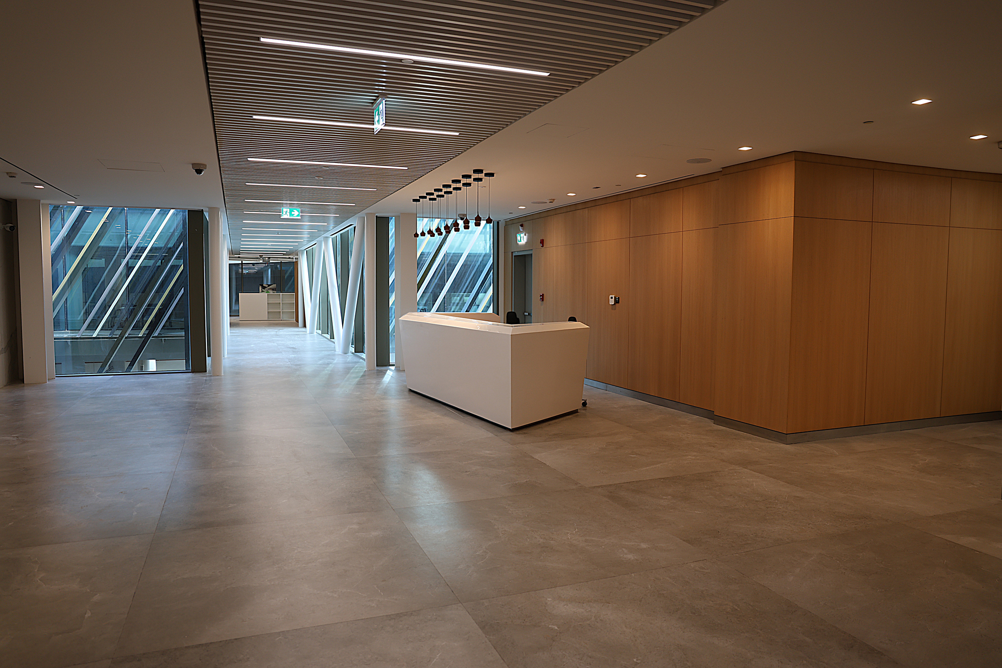 Reception lobby with white desk, wood paneling and slatted ceiling beside glazed facade at evening