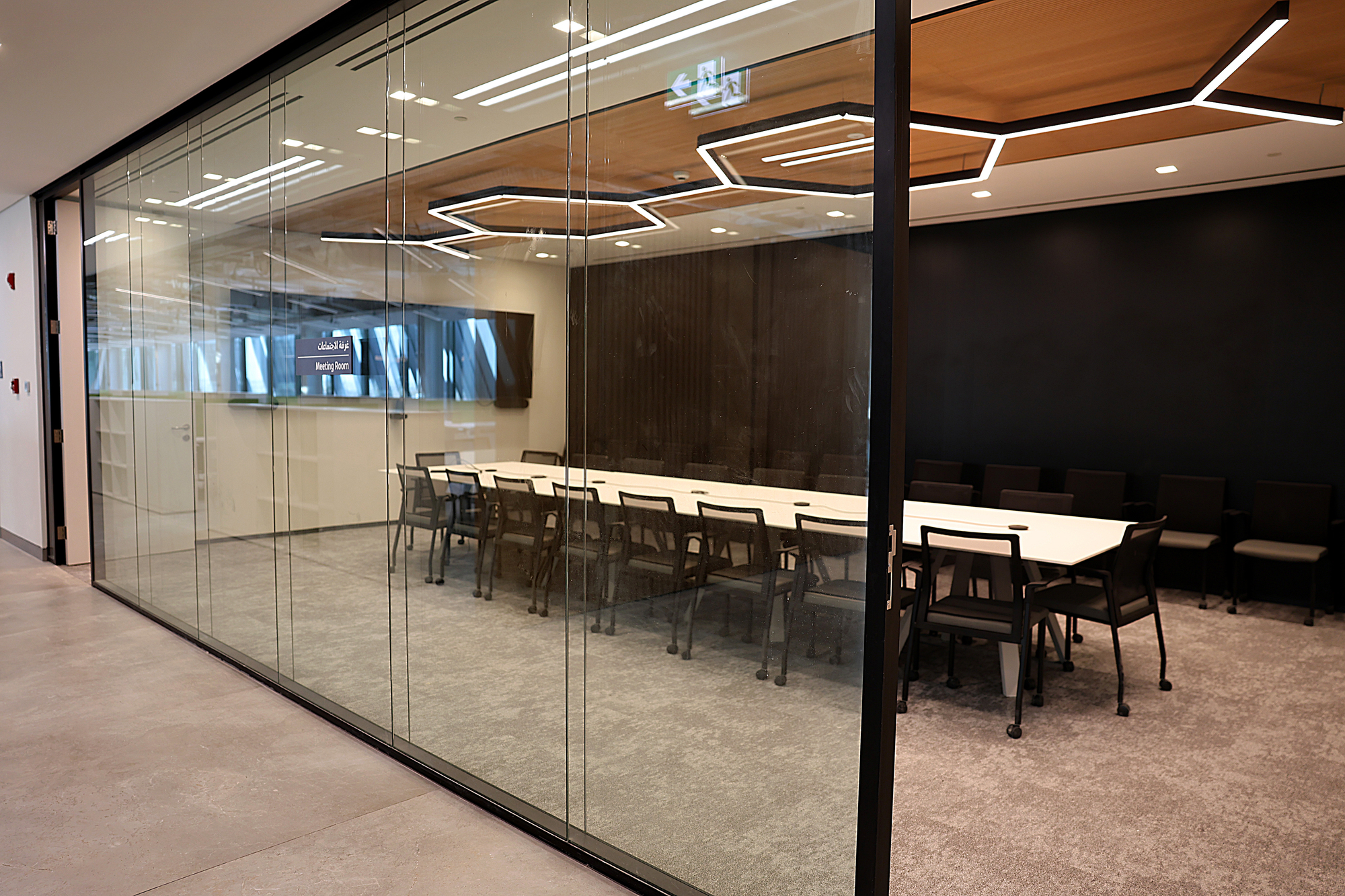 Glass-walled conference room with long table, dark chairs and hexagonal ceiling light fixtures viewed from corridor