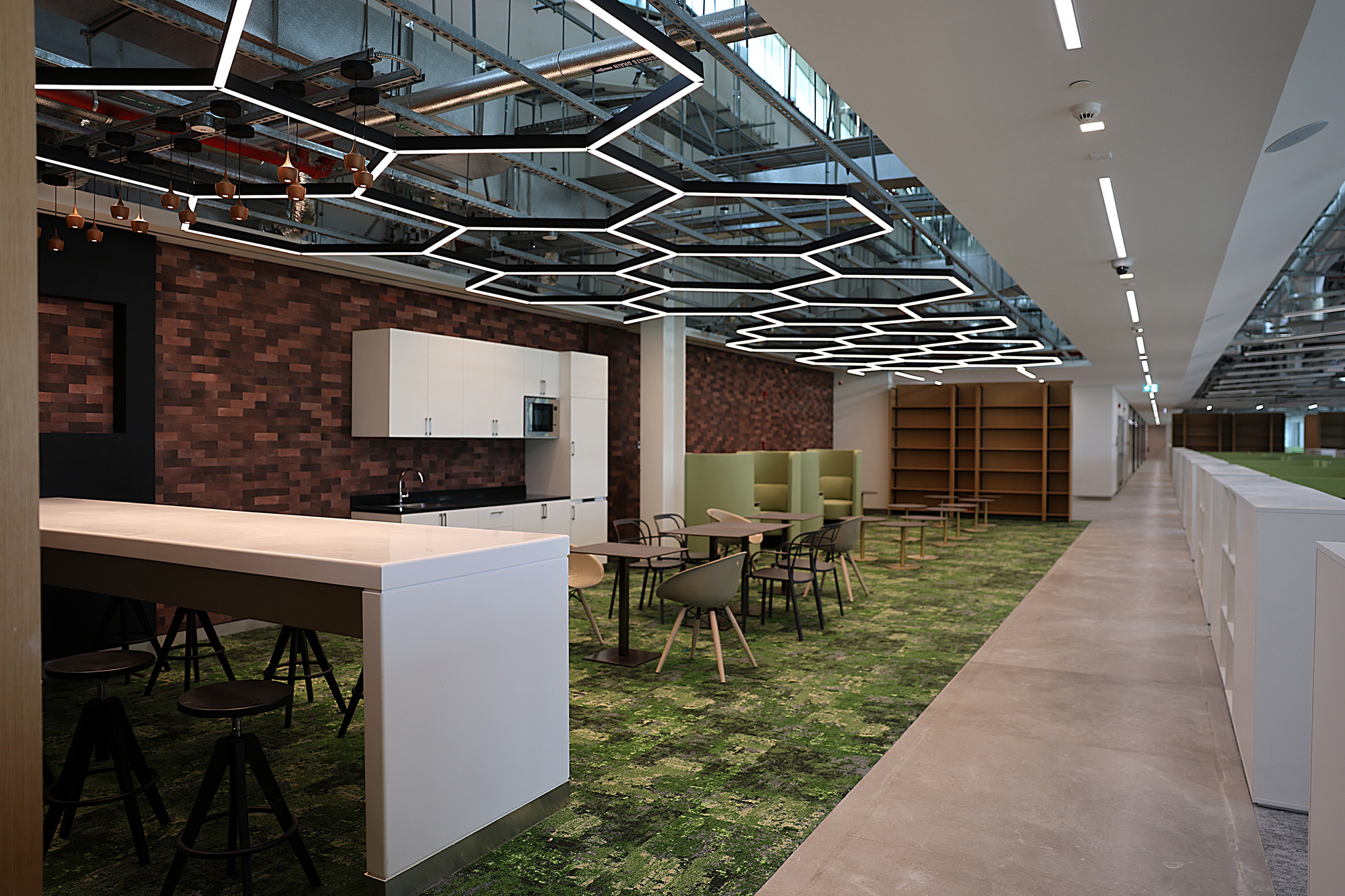 Breakout area with white kitchen island, grass-patterned carpet and geometric lighting overhead with exposed mezzanine