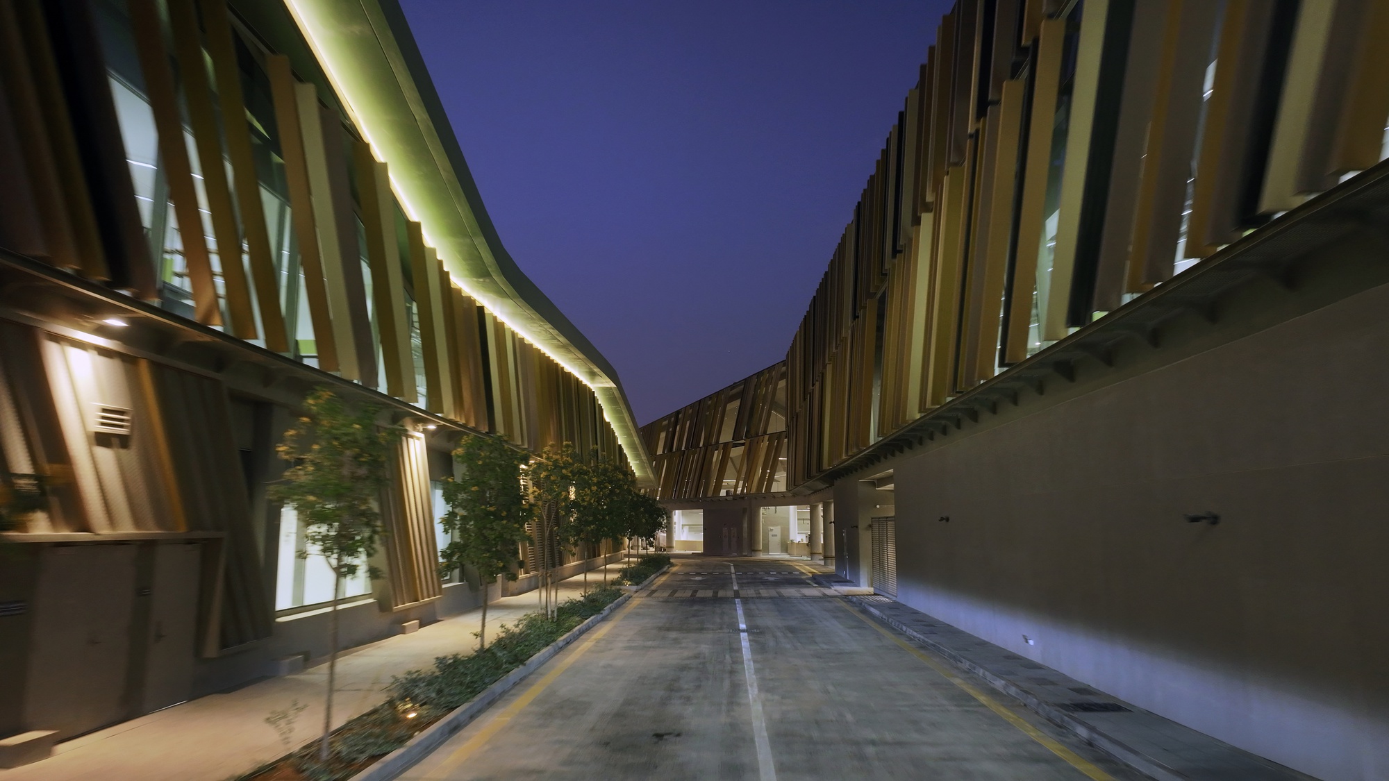 Illuminated pedestrian passage between vertical metal screen facades and planted median at dusk