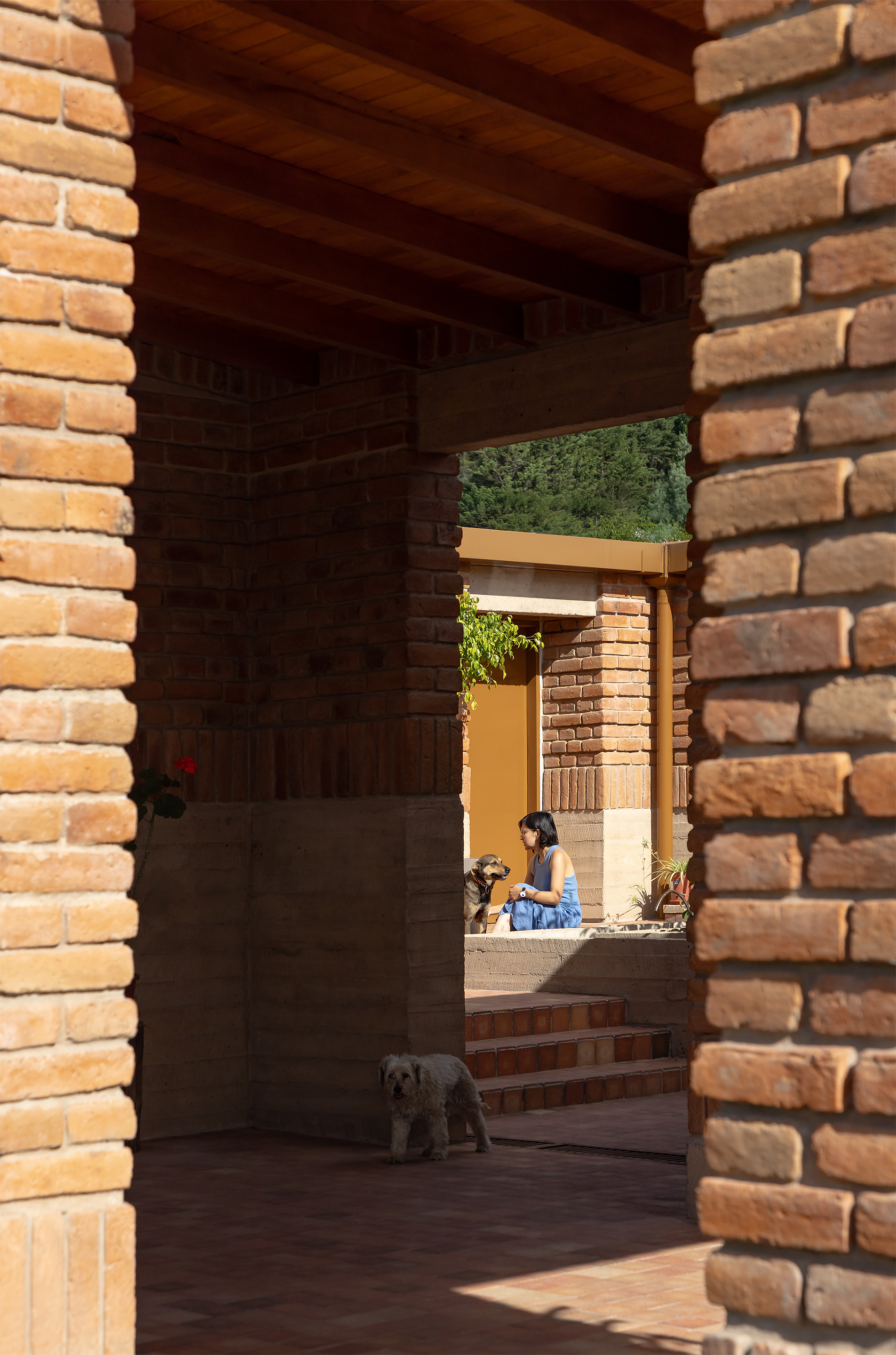 Shaded brick corridor with timber ceiling beams framing a view of a seated figure and dog