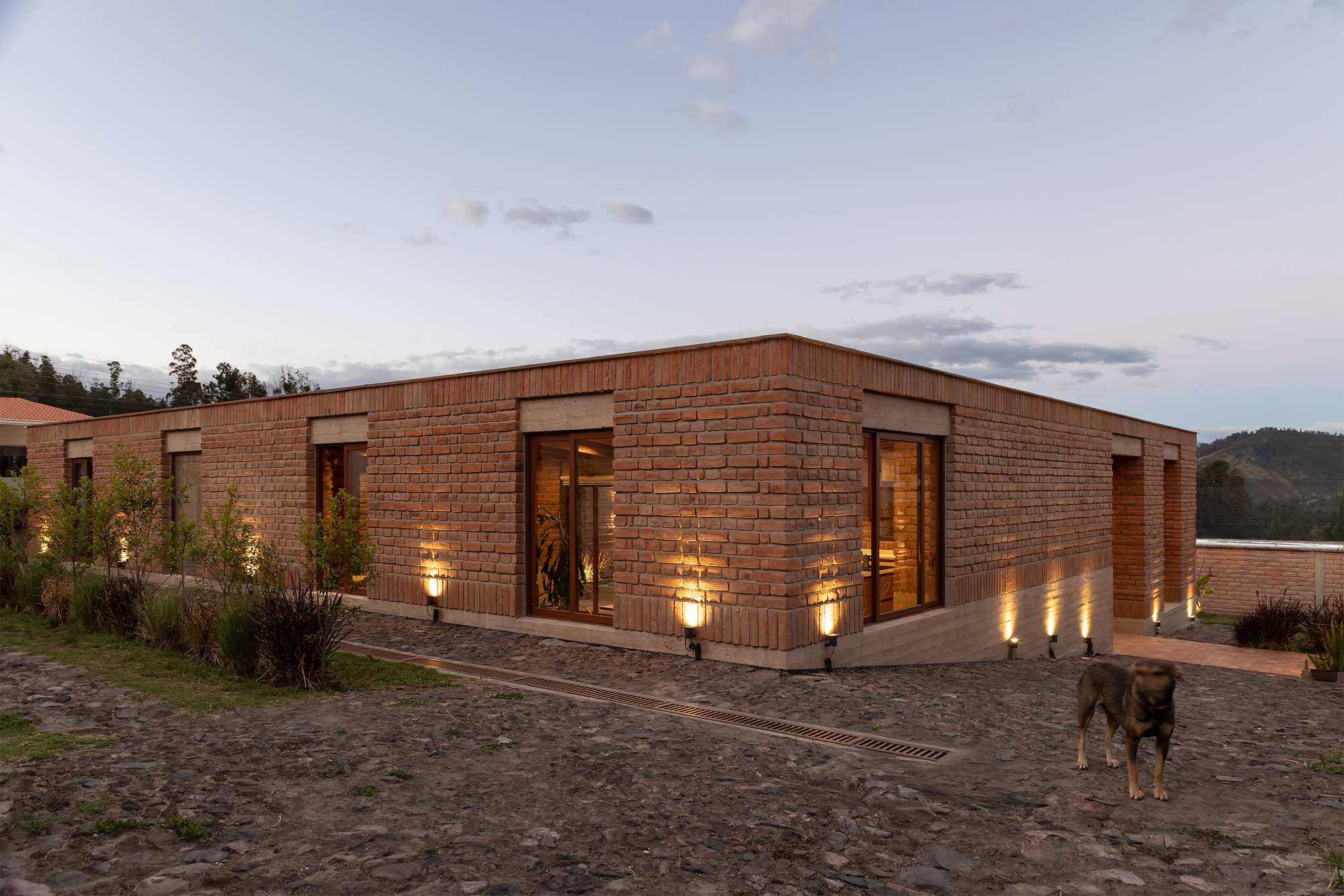 Corner view of the brick volume with uplighting and a dog on the cobbled drive