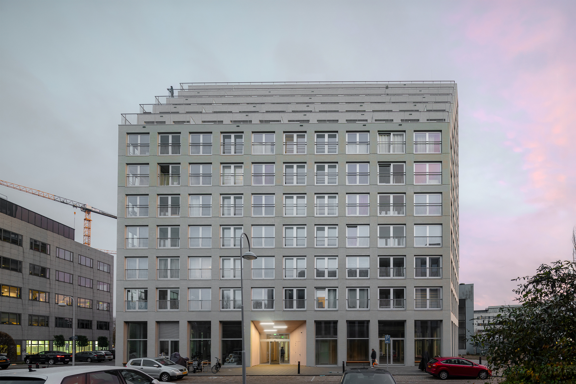 Street view of the gridded facade with rooftop pergola structure at dusk under pink sky