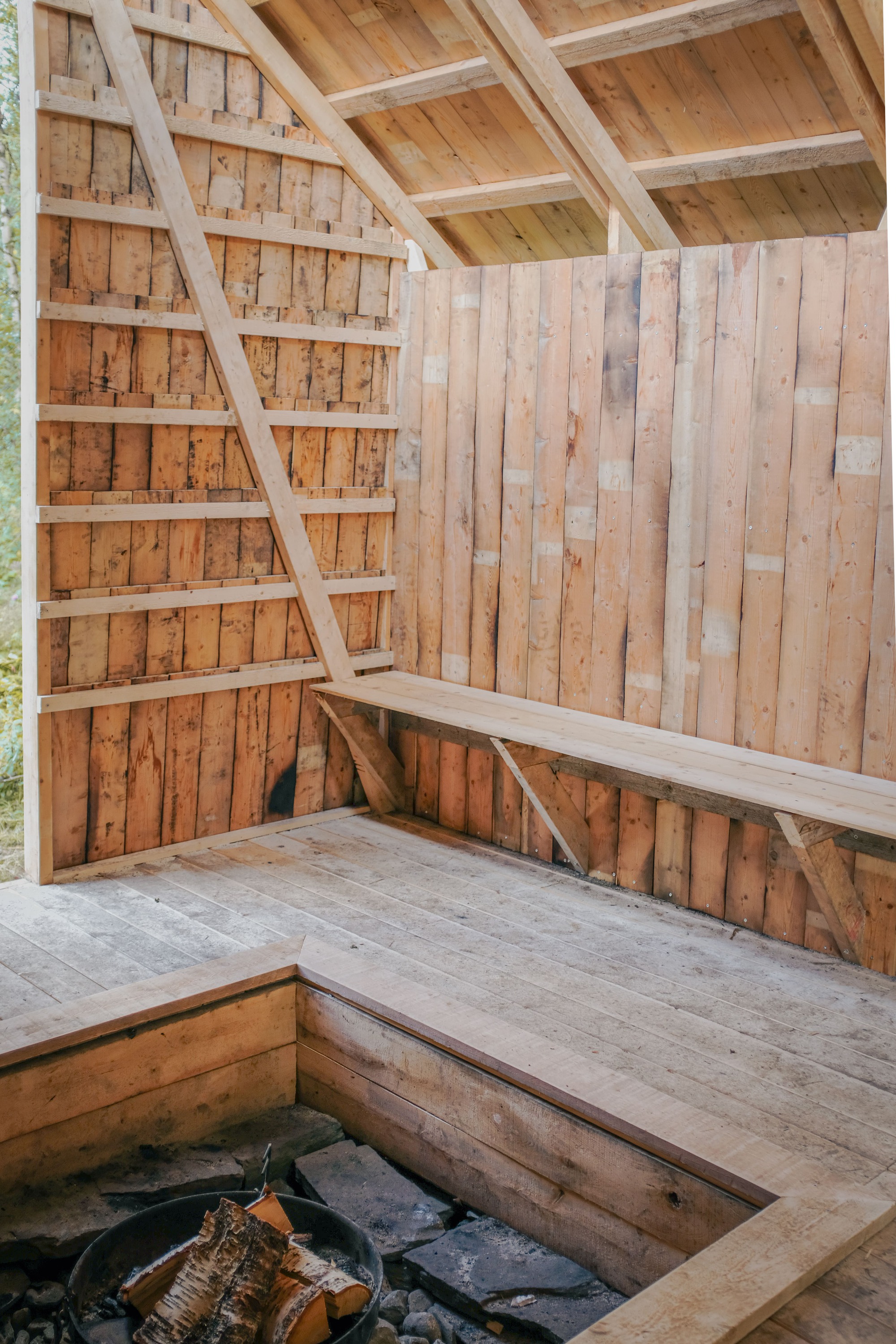 Corner of timber sauna interior with diagonal bracing, benches, and fire pit opening