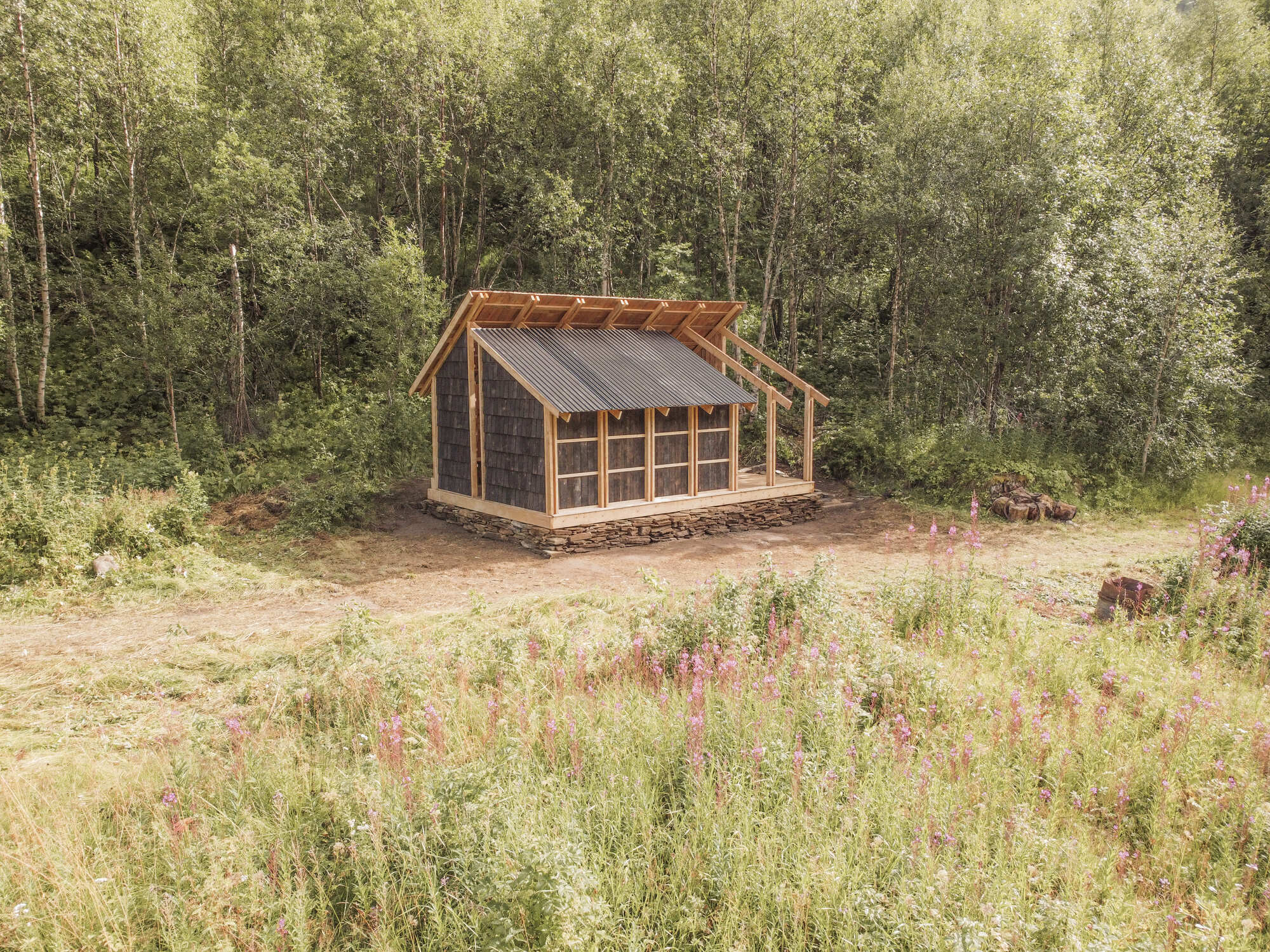 Gabled timber pavilion with stone base and metal roof in a forest clearing