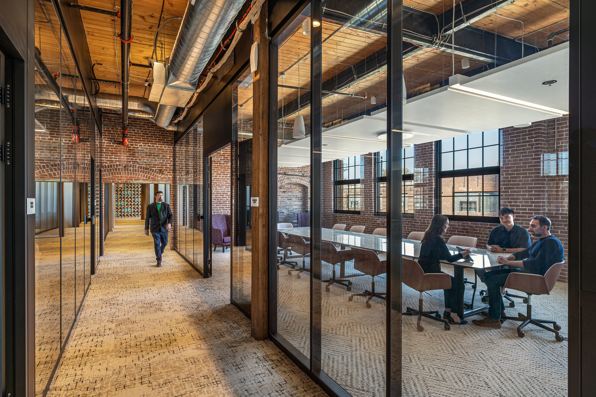 Glazed corridor with black steel frames alongside a conference room with exposed brick walls